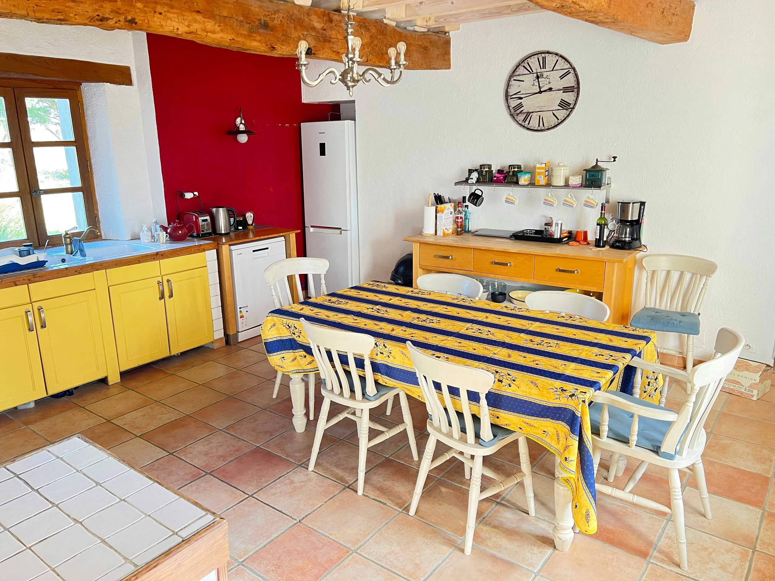 A kitchen and dining area with yellow cabinets, a red accent wall, a wooden ceiling, a chandelier, and a large clock on the wall. There is a blue and yellow tablecloth on the dining table with white chairs around it. Various kitchen appliances and utensils are on the countertop.