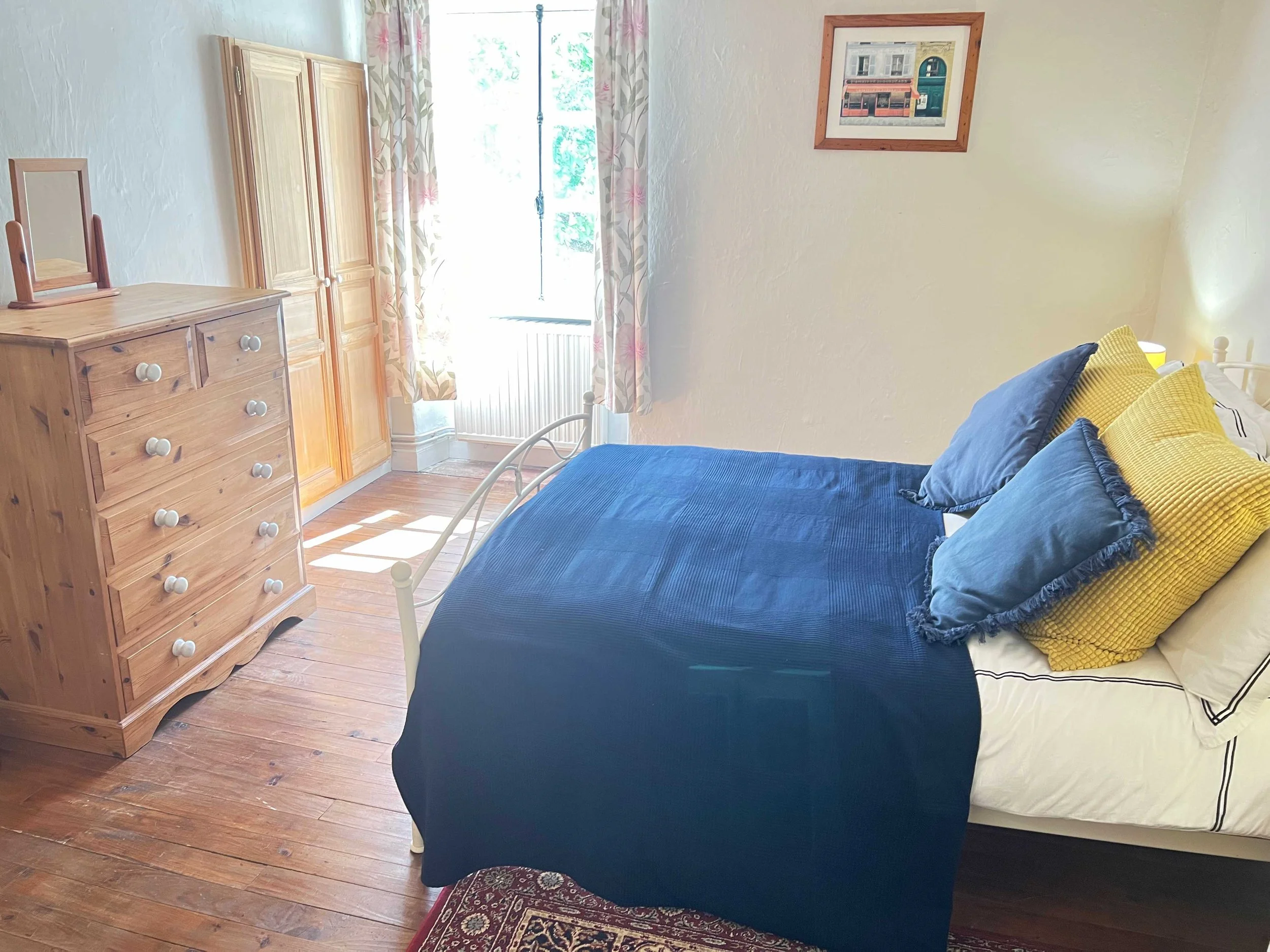 A bedroom with a wooden dresser, a wardrobe, a bed with blue bedsheets and yellow pillows, a window with floral curtains, a framed picture on the wall, and hardwood flooring.