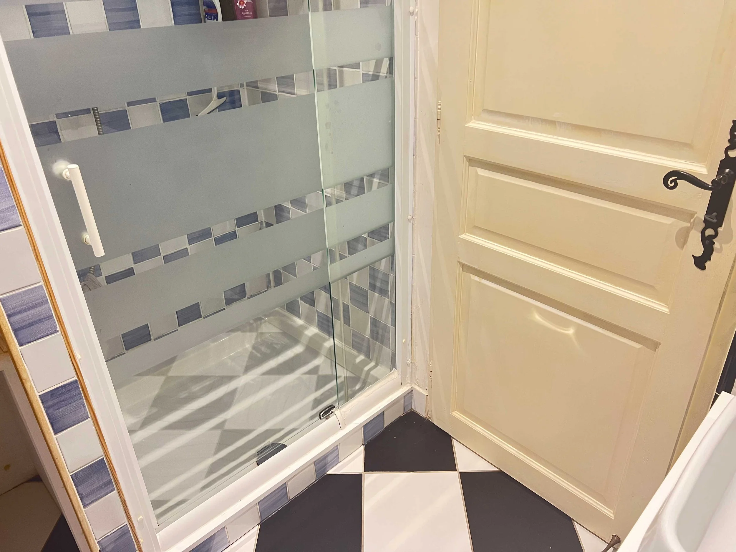 A door with a black decorative handle and paneled design next to a glass shower enclosure with frosted horizontal stripes and blue tile accents in the bathroom. The bathroom has black and white checkered floor tiles.