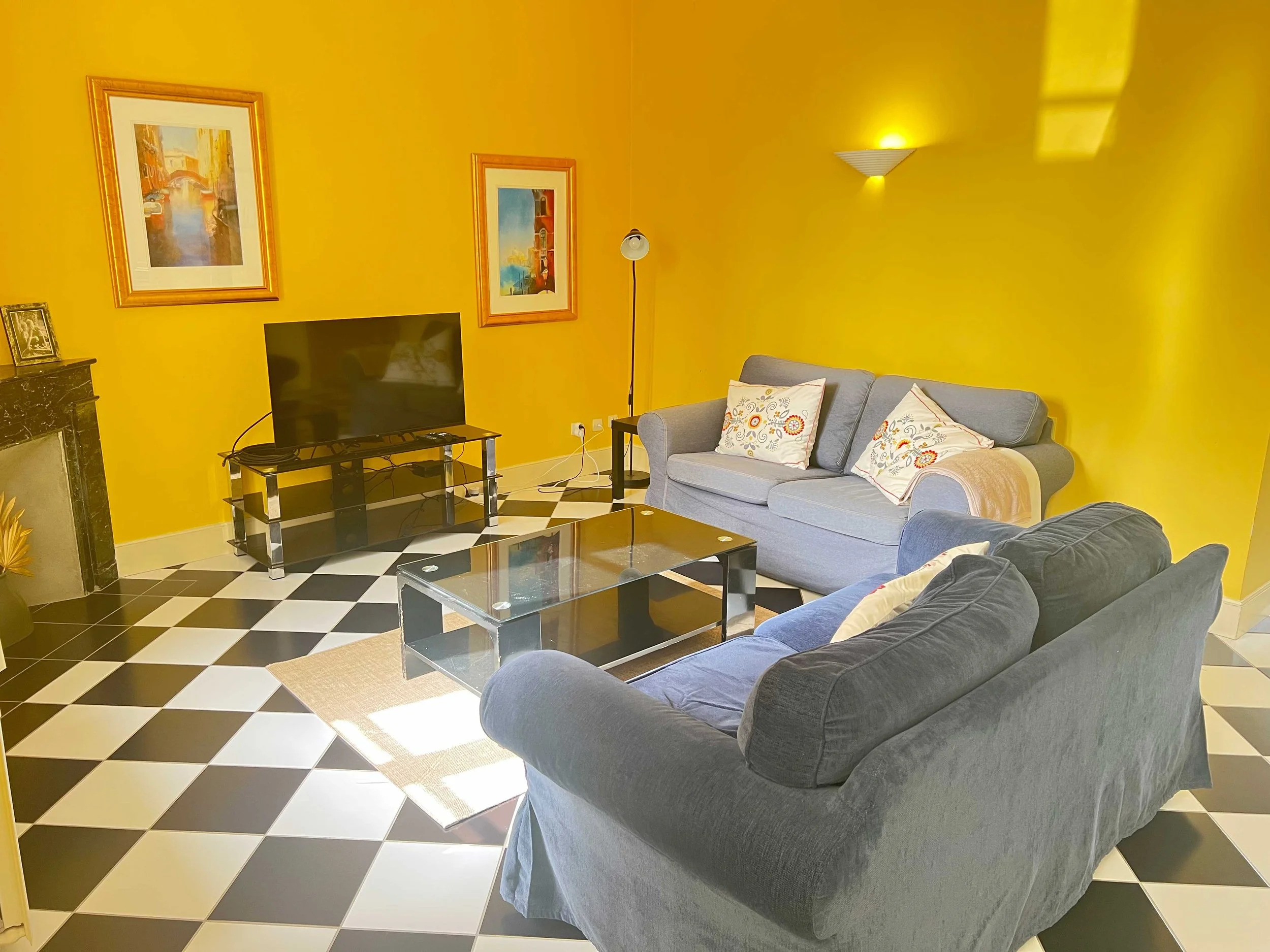 Living room with yellow walls, a gray sofa with decorative pillows, a glass coffee table, a black entertainment center with a TV, framed artwork on the walls, a floor lamp, and a checkerboard black and white tile floor.