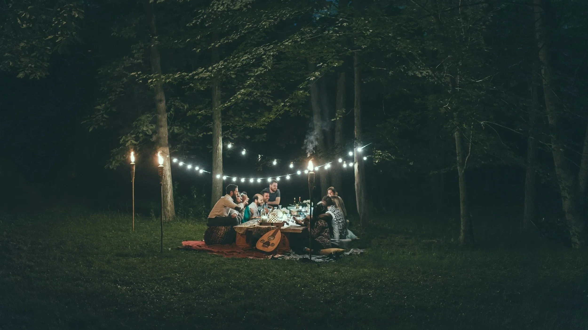 A group of people enjoying a night gathering around a table outdoors in a wooded area, lit by string lights and torches, with some playing music instruments.