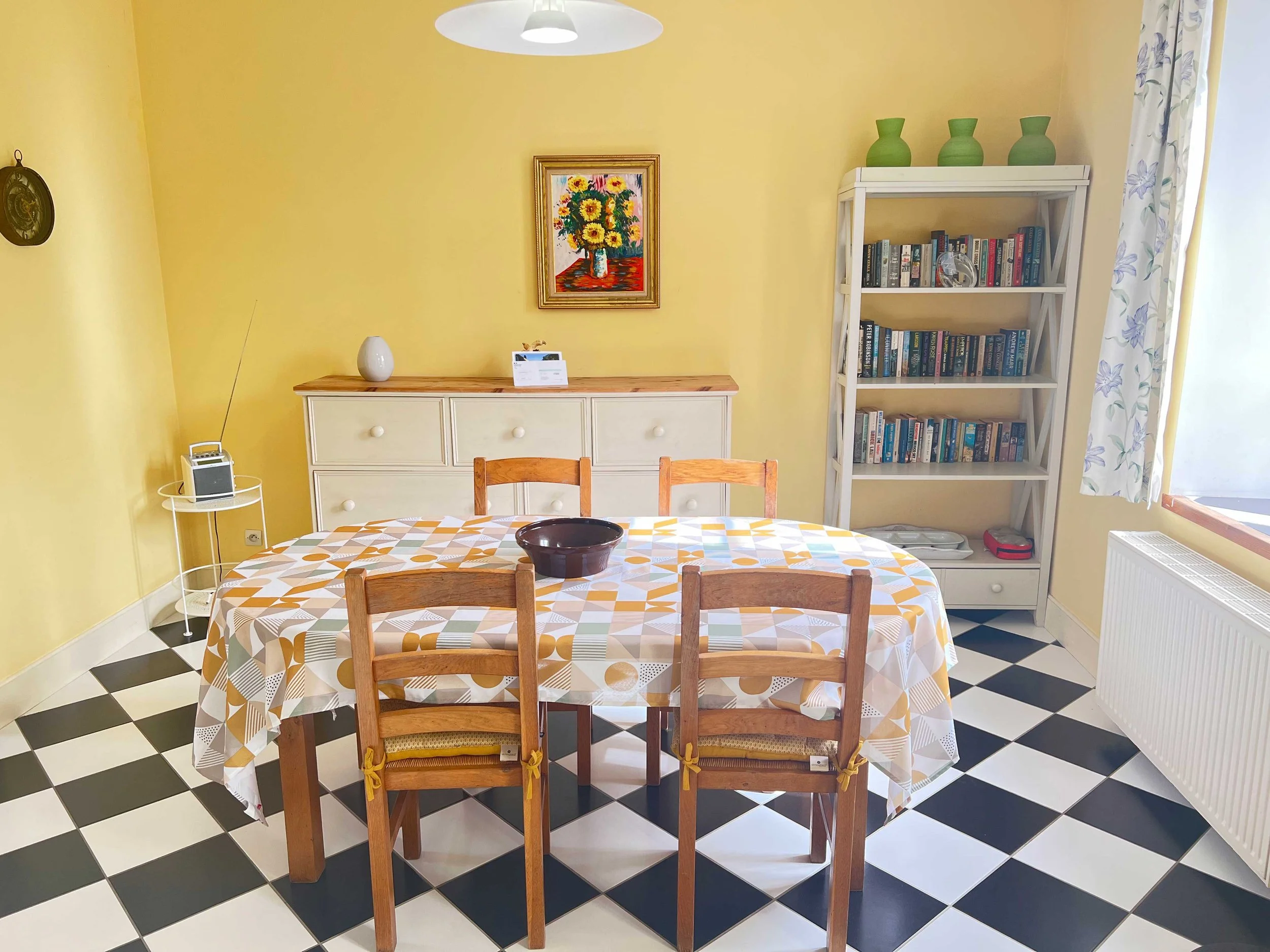 A yellow-dappled wall with a framed painting of sunflowers, a white sideboard with a white vase, a black bowl on a table with covered chairs, a bookshelf with green vases on top, and a window with floral curtains.