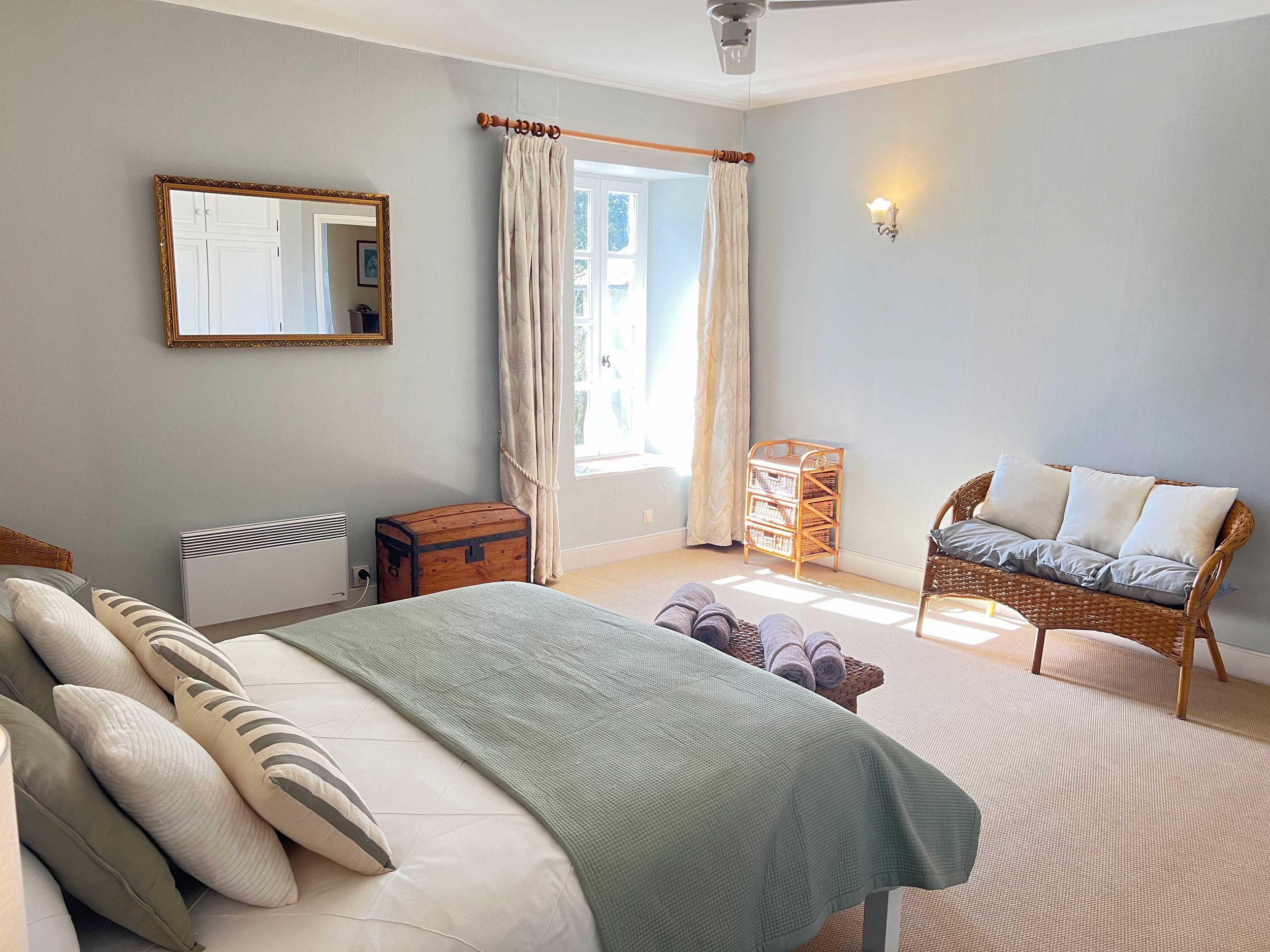 A bedroom with a bed covered in light green bedding and multiple pillows, a wicker chair with a white cushion, a small wooden chest, a window with curtains letting in natural light, a wall sconce, and a mirror on the wall.