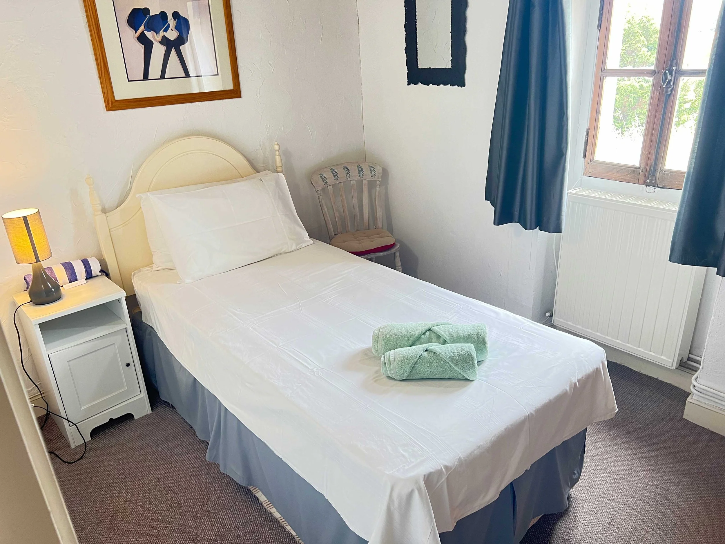 A cozy bedroom with a white bed, green towels on top, a side table with a lamp and striped towel, a wooden chair with a cushion, a window with blue curtains and a view of green trees outside, and framed artwork on the wall.