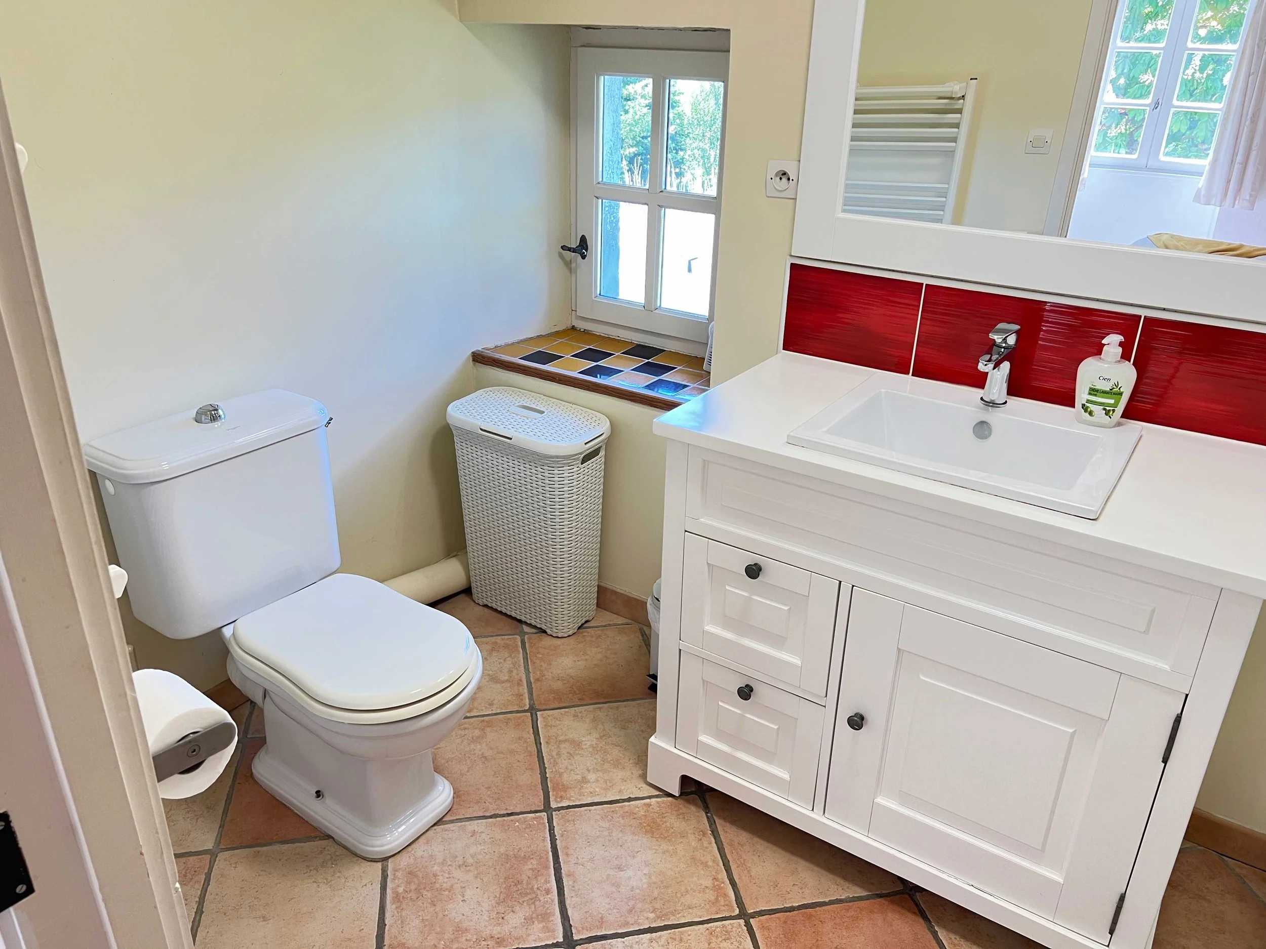 A small bathroom with a white toilet, a white wicker laundry basket, a white sink with a dark red backsplash, a mirror, a towel warmer, and a window with sunlight and trees outside.