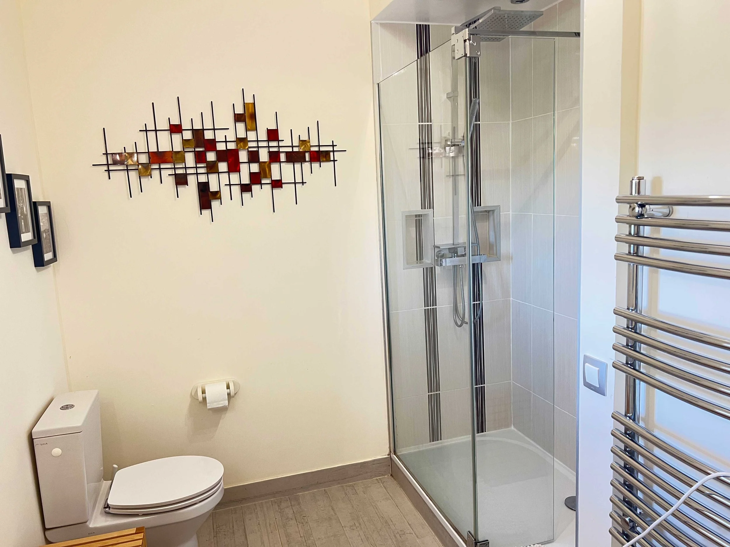 A bathroom with a toilet, a glass shower enclosure, a wall-mounted shower head, a heated towel rail, and wall decor including framed pictures and a metal art piece.