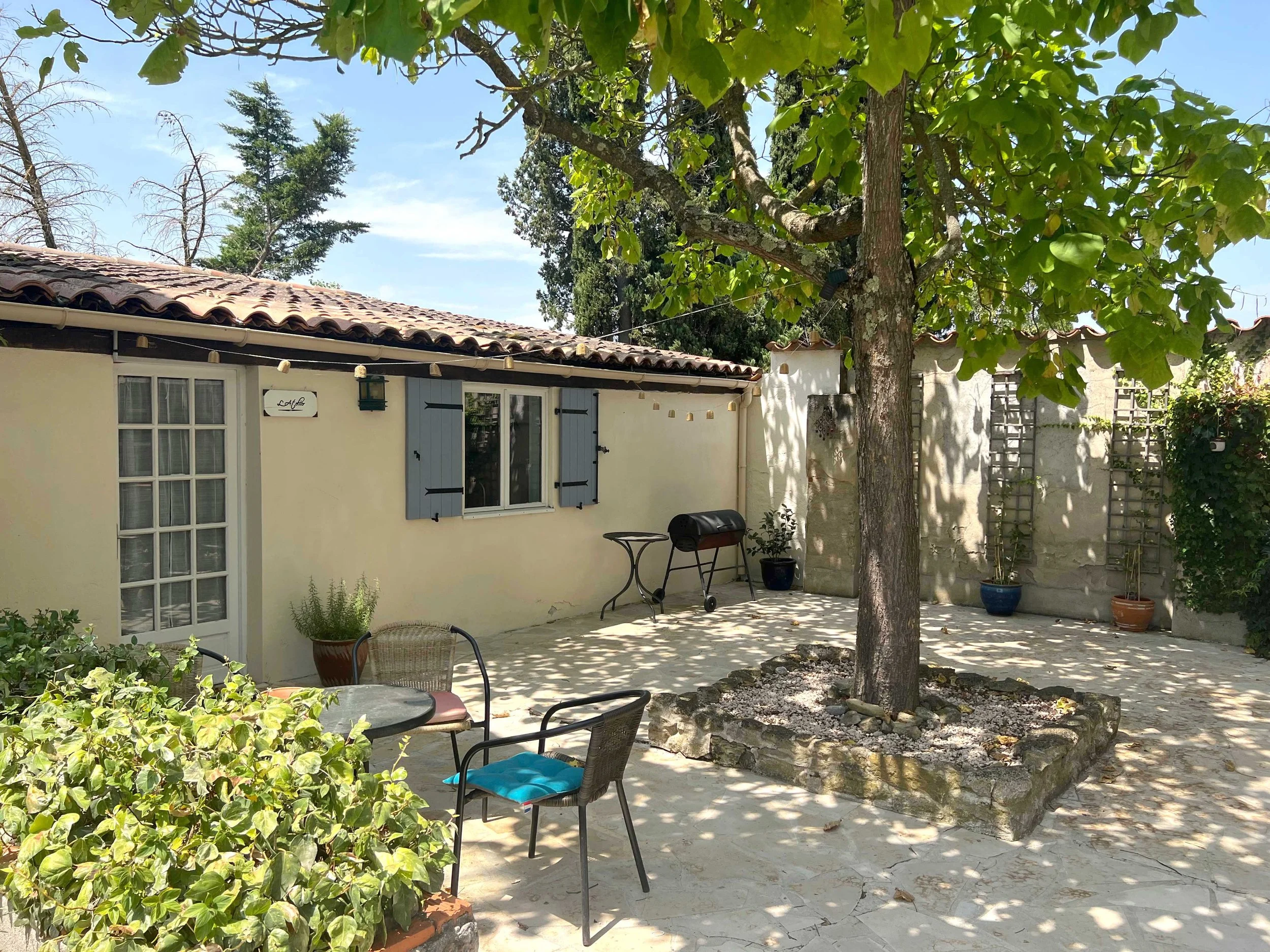 A backyard patio featuring an outdoor table with four chairs, a small grill, potted plants, and a large tree with dense green foliage providing shade. The patio is adjacent to a house with cream-colored walls, blue shutters, and a tiled roof. There are string lights hanging above and a wooden trellis on the right side.
