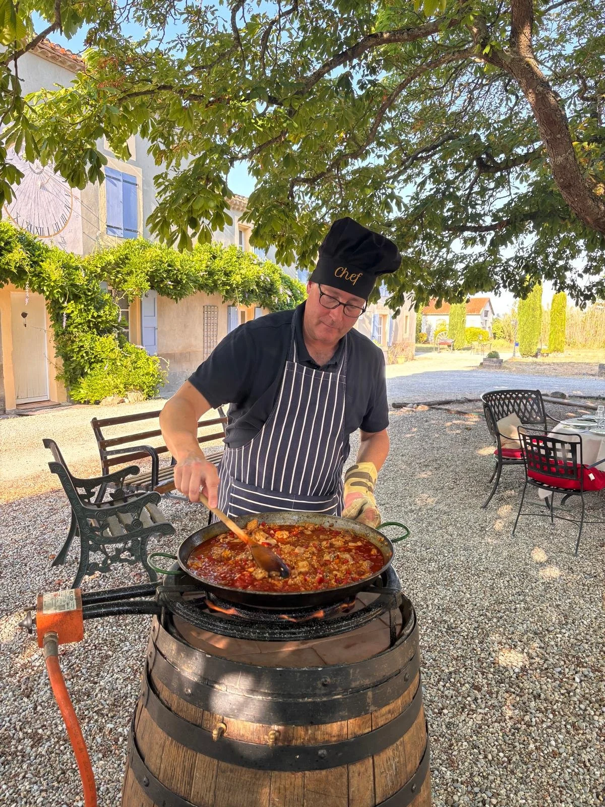 A man wearing sunglasses and a black apron stands outdoors near a barrel grill, cooking a colorful vegetable dish. The background includes tall green trees, a stone patio, and outdoor furniture under a clear blue sky.