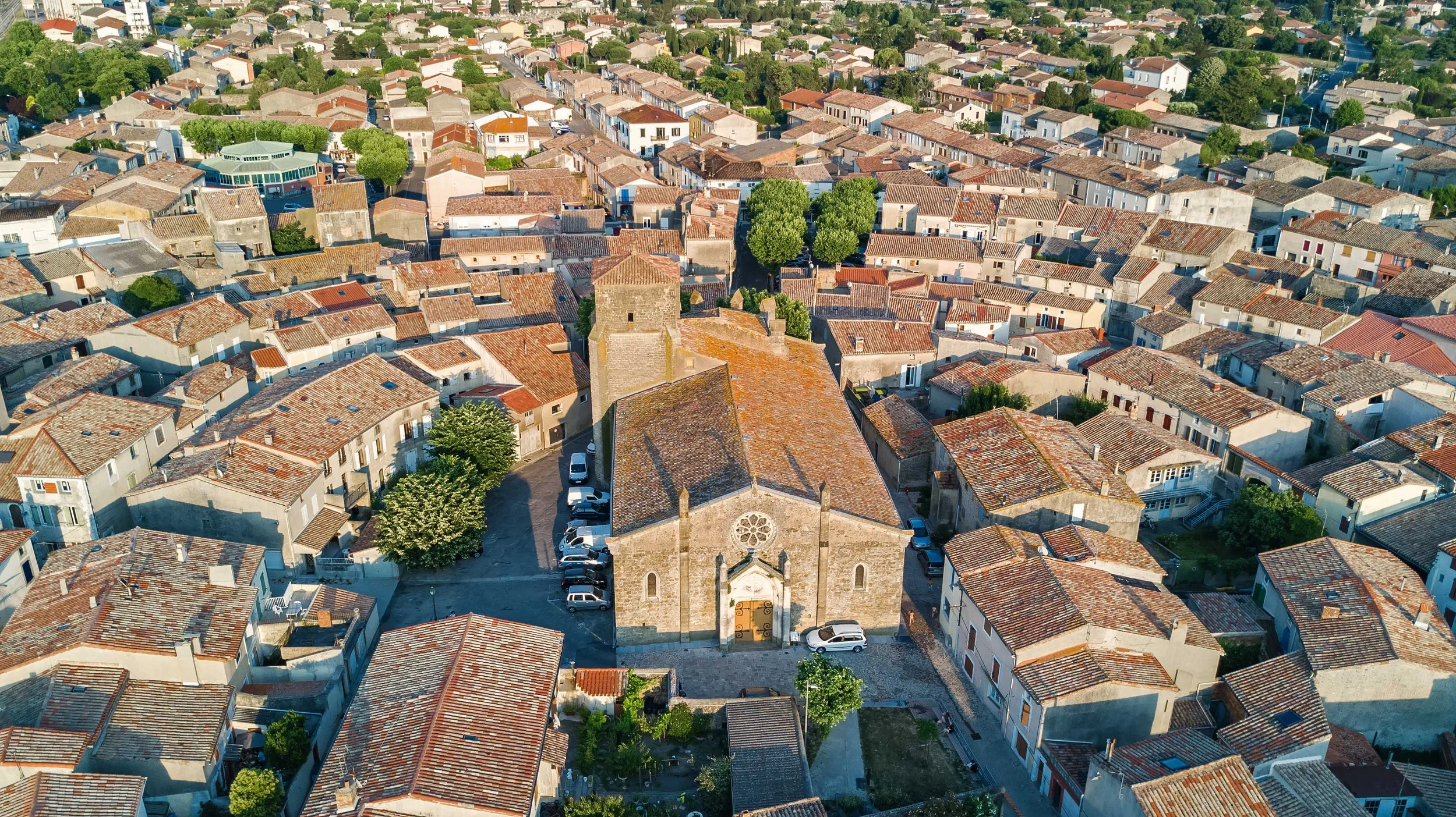 Aerial view of a European town showing tightly packed houses with red-tile roofs, a historic stone church with a tower, and several trees among the buildings.