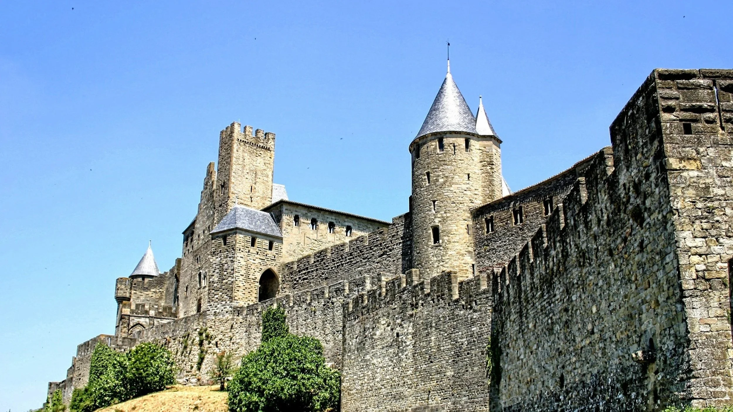 A medieval castle with stone walls, towers, and conical rooftops under a clear blue sky.
