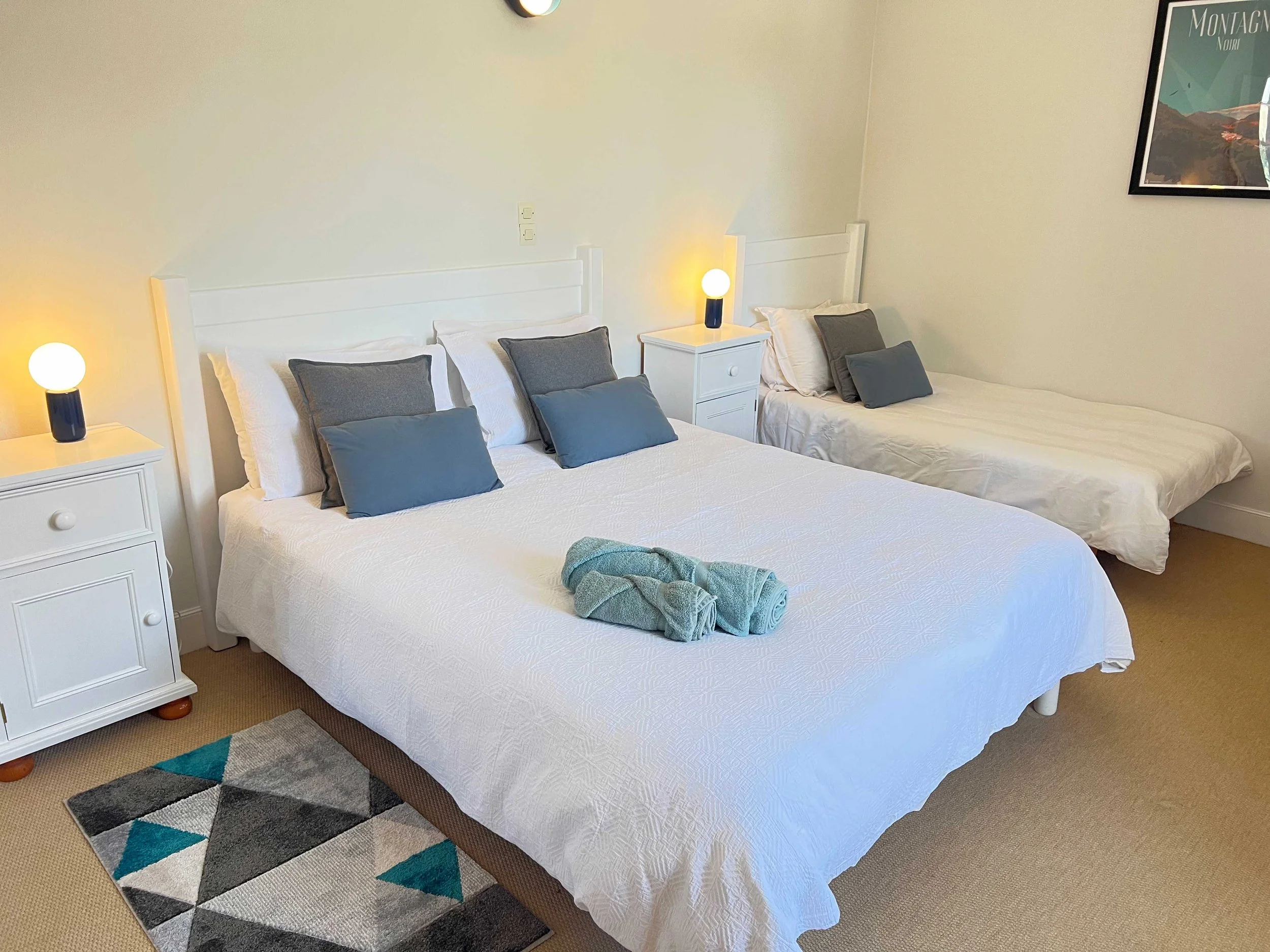 A bedroom with two white beds, each with white bedding and gray pillows. There are nightstands with lamps on each side of the beds, and a framed poster on the wall. A small colorful rug is on the beige carpet. A small towel is folded on the bed in the foreground.