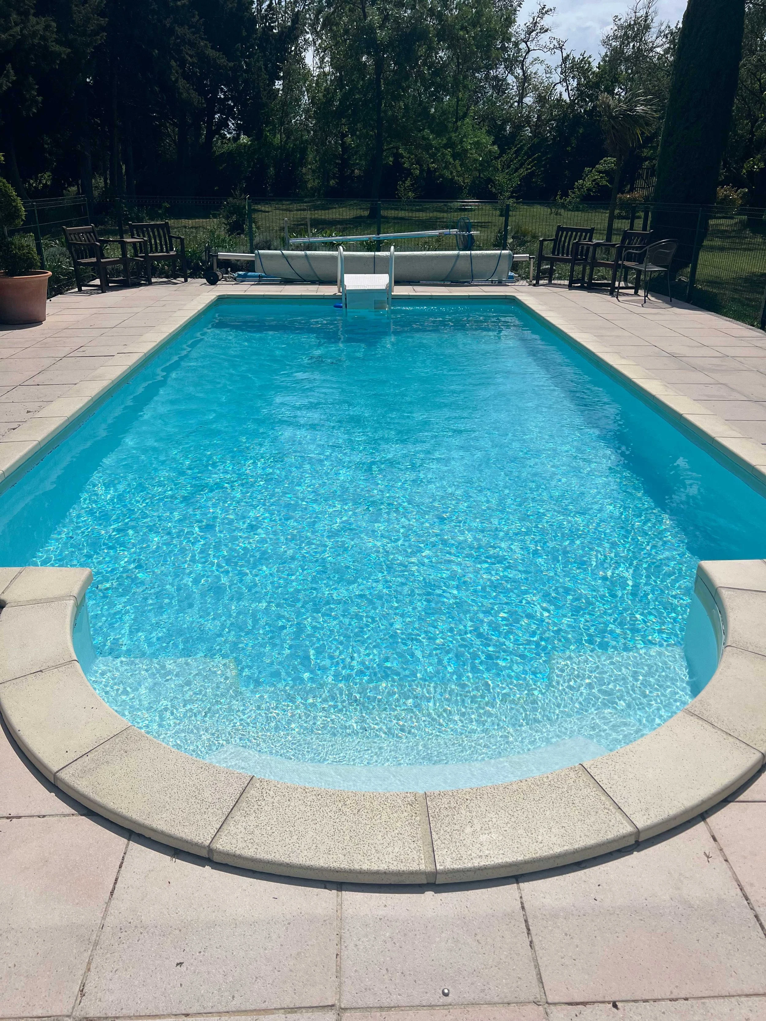 A backyard swimming pool with clear blue water, surrounded by a light-colored tiled deck, with a small set of steps leading into the pool. In the background, there are several black chairs, some with armrests, arranged along the fence, and a large tree with lush green foliage providing some shade. There are also some bushes and trees beyond the fence.