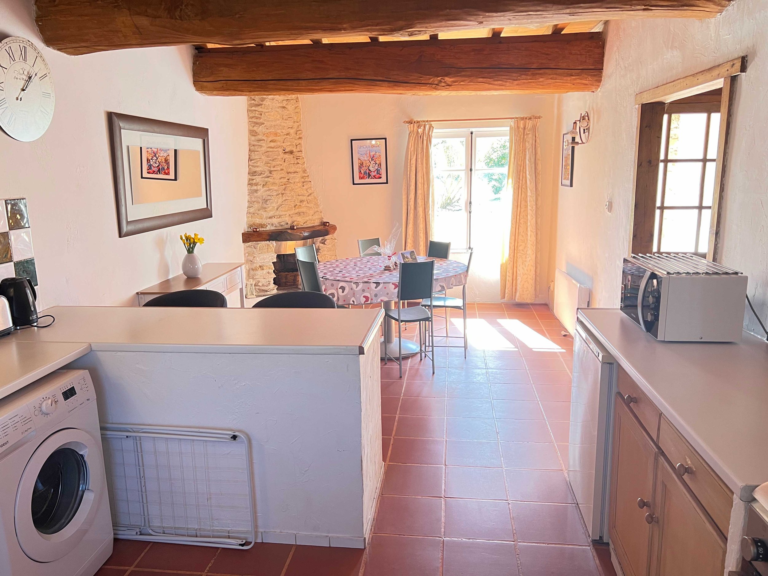 A kitchen and dining area with a round table covered with a colorful polka dot tablecloth, four chairs, a window with yellow curtains, a shelf with picture frames, a stone and wood fireplace, and a washer and microwave visible in the kitchen area.
