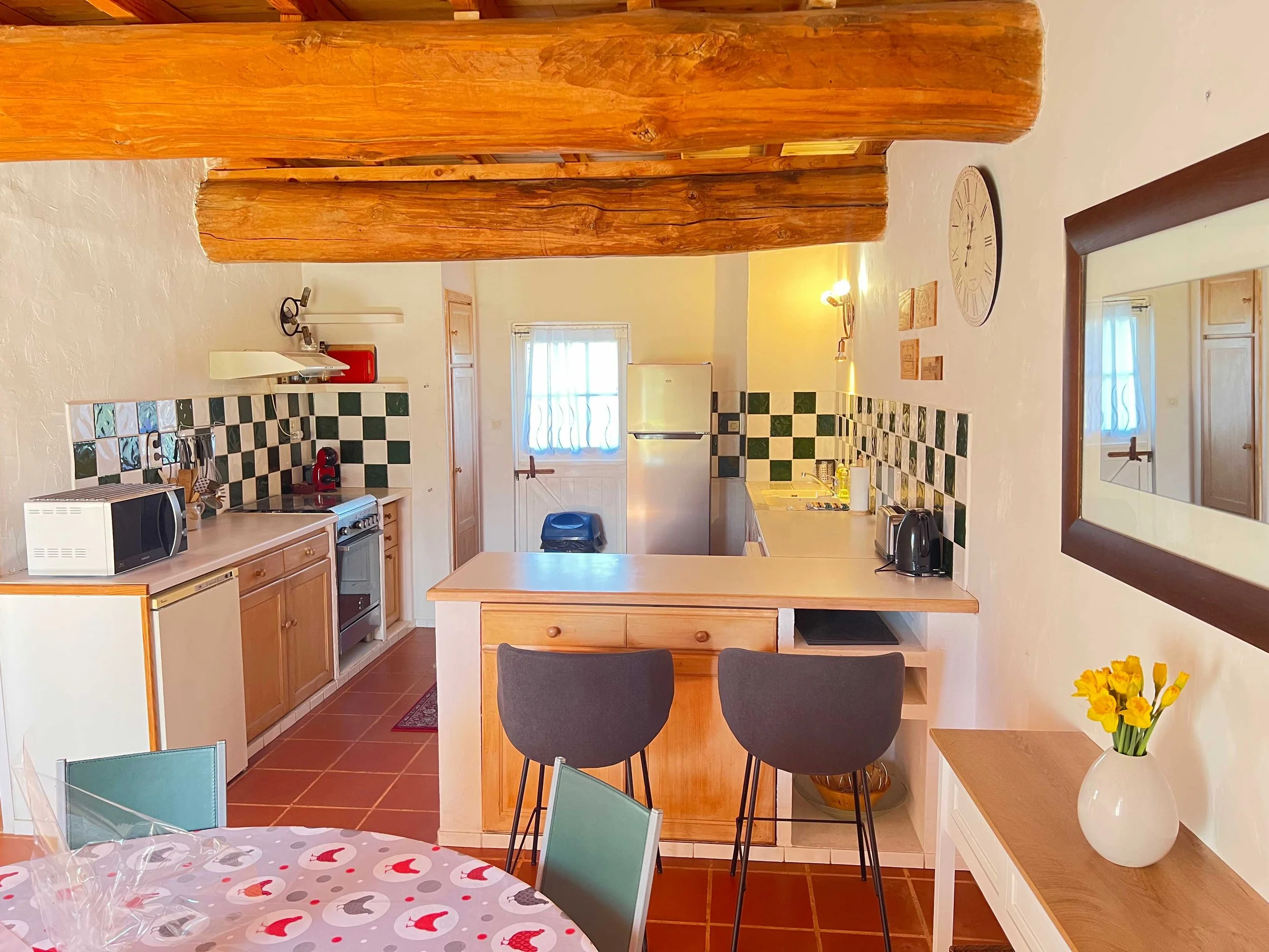 A cozy kitchen with wooden beams, green and white checkered backsplash, white appliances, and a dining area with gray chairs and a table with a festive tablecloth featuring red and gray foxes. There is a vase of yellow flowers on a side table.