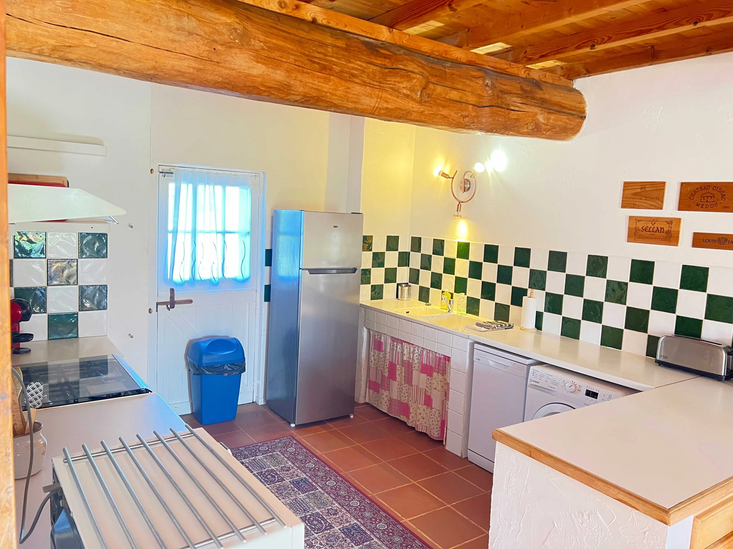 A cozy kitchen with a window, a refrigerator, a sink with a floral curtain underneath, and a washing machine. The kitchen has green and white checkered tiles on the wall, wooden beams on the ceiling, and a small rug on the terracotta tile floor.