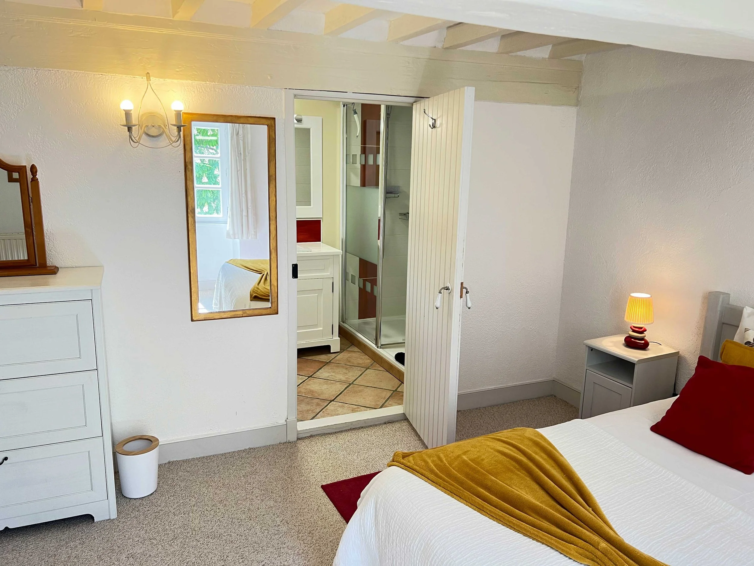 A cozy bedroom with a bed dressed in white linens, accented with a red pillow and a mustard-colored blanket. There is a small white nightstand with a colorful lamp on it, and a dresser with a mirror on the left. A wall-mounted light fixture illuminates the room. A sliding door leads to a bathroom with a shower enclosure, a small window, and a cabinet.