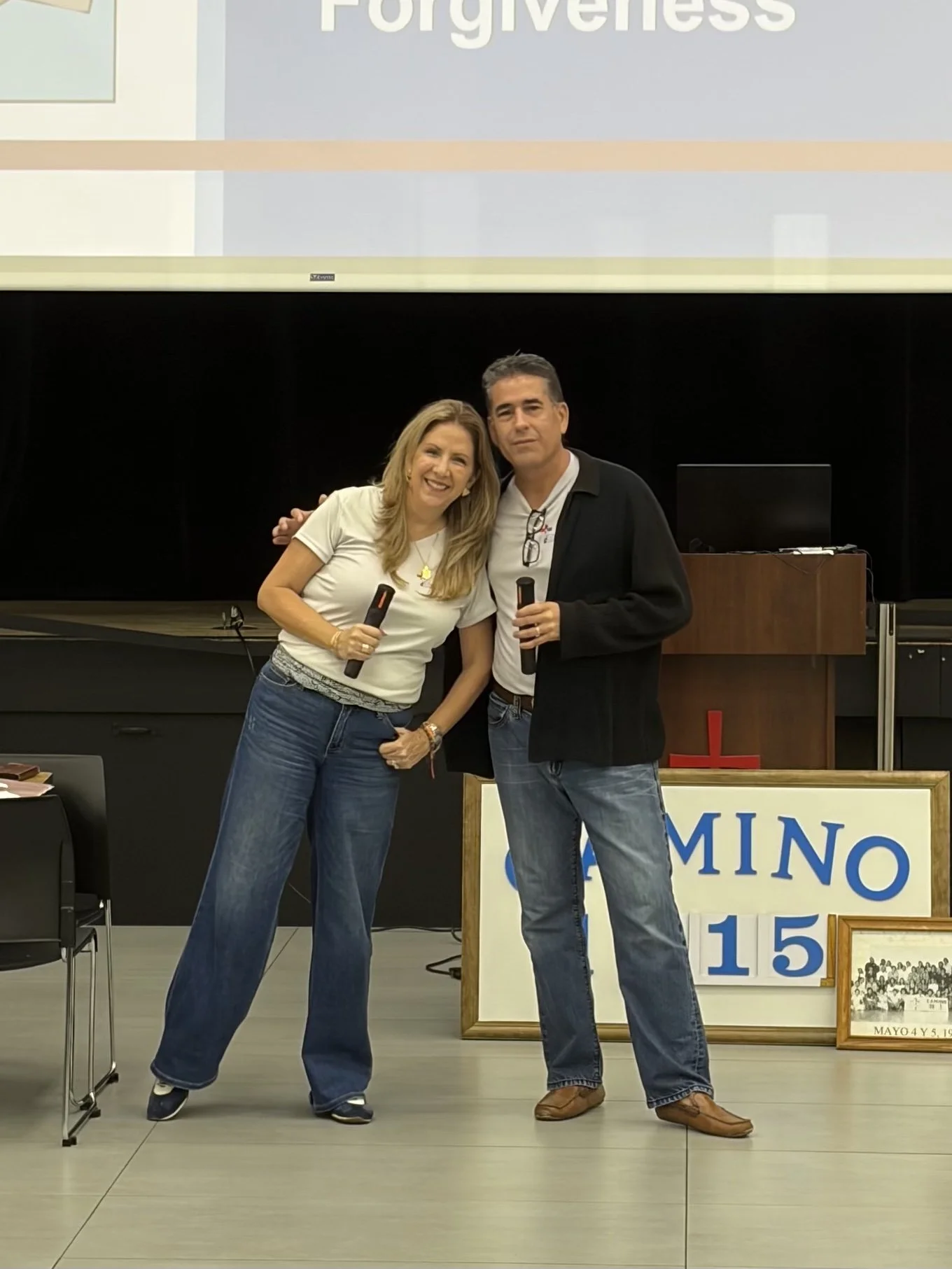 Two people, a woman and a man, smiling and holding microphones, standing in front of a presentation screen and a sign that reads 'Mino 15'. The woman has long blonde hair and is wearing a white t-shirt and blue jeans. The man has short dark hair and is wearing a white shirt, black jacket, and blue jeans.