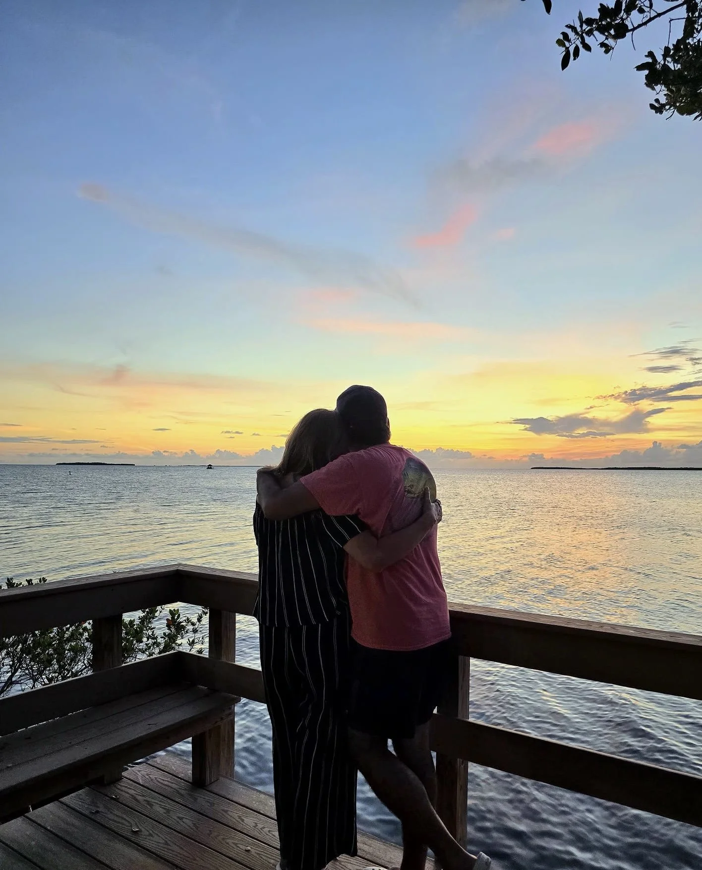 A couple hugging on a wooden deck overlooking a body of water at sunset, with colorful sky and clouds in the background.