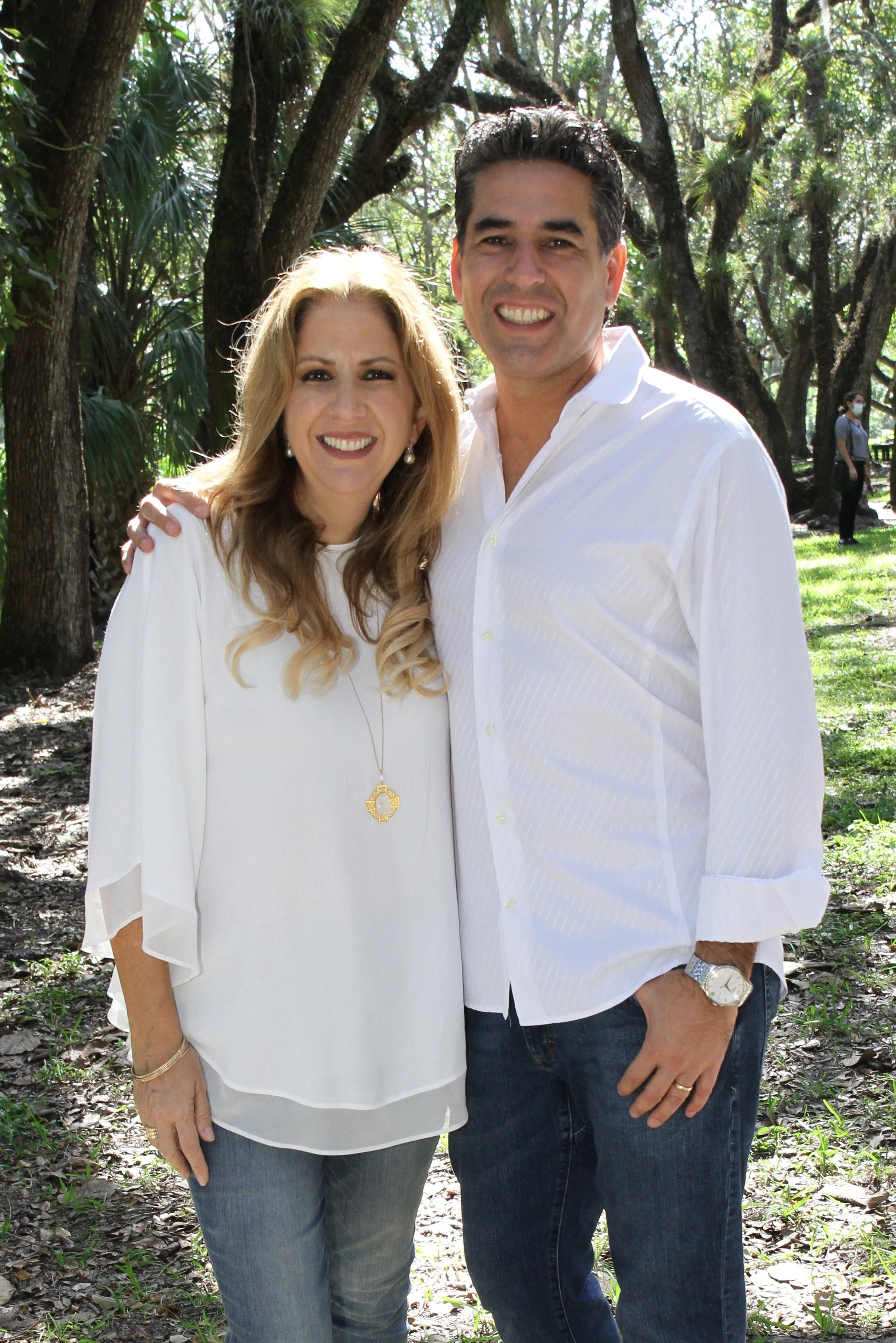 A smiling middle-aged woman with blonde hair, earrings, and a white blouse standing next to a smiling middle-aged man with dark hair, wearing a white shirt and blue jeans, in a park with trees and sunlight.