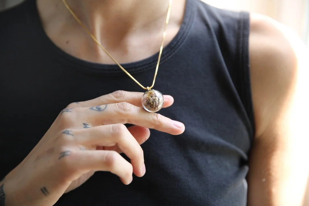 Close-up of a woman wearing a black sleeveless top and a delicate gold necklace with a round pendant containing small flowers.
