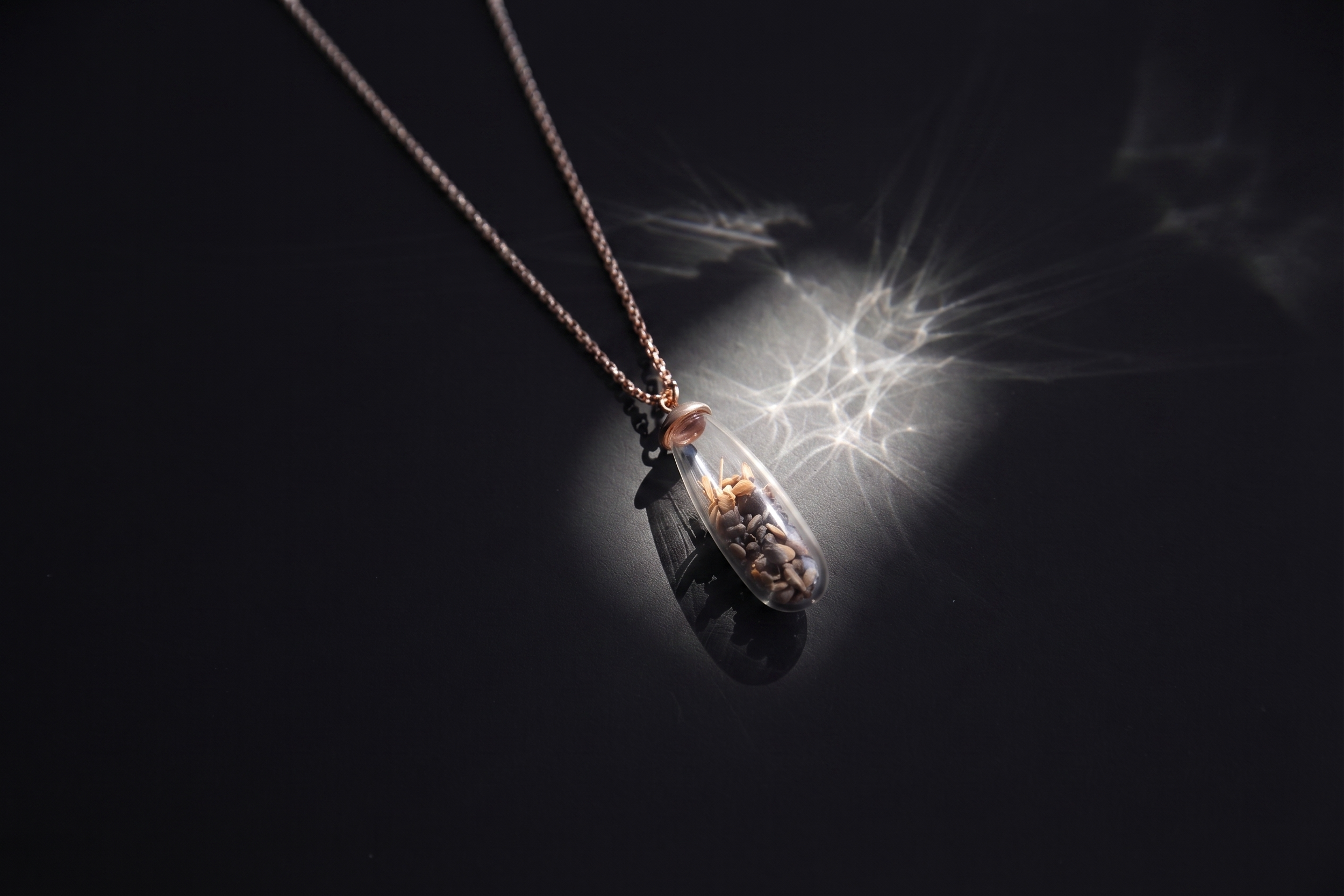 A necklace with a glass pendant containing small rocks and dried flowers, casting a star-shaped light and shadow on a black surface.