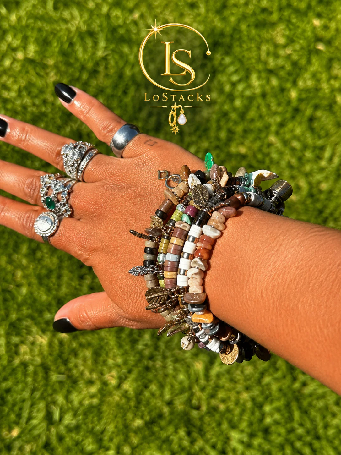 A hand with multiple silver rings and black nail polish, wearing a stack of colorful beaded bracelets with leaf charms. The background is green grass, and there is a logo for LoftStack in the upper part of the image.