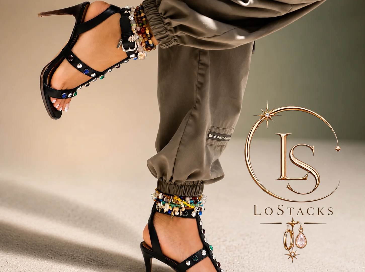 Close-up of a model's legs and feet showing embellished high-heeled sandals with colorful beads and charms, khaki cargo pants with zipper detail, and a silver and gold jewelry stack with charms. A logo with the initials L S and the text 'LoStacks' with star and celestial motif is also present.