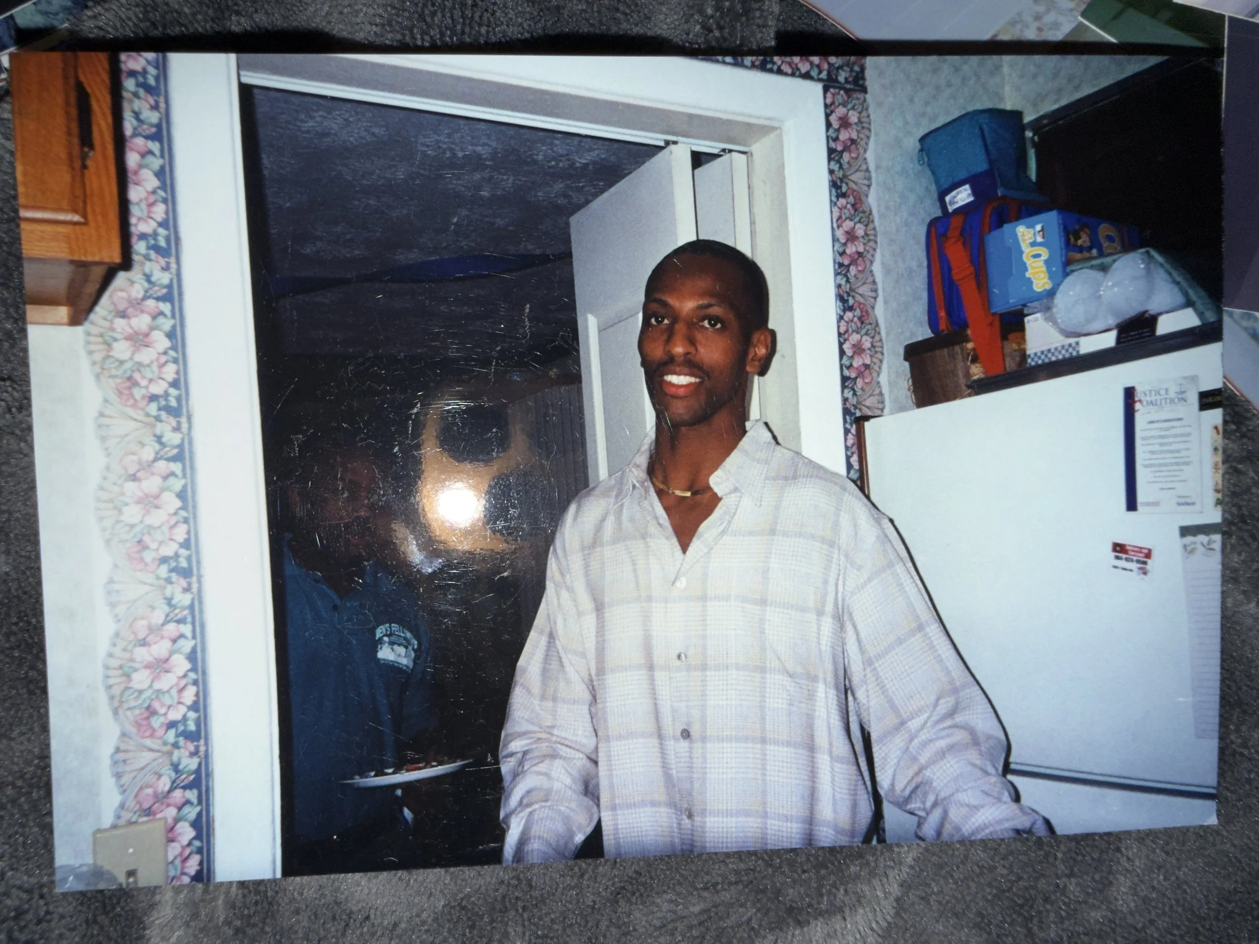 A man with dark skin, short hair, wearing a light checkered shirt, standing in a kitchen in front of a window with floral curtains.