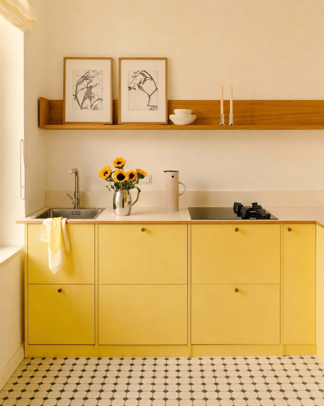 Yellow kitchen cabinet with a vase of sunflowers, a beige kettle, black cooktop, sink, and two framed botanical prints on a wooden shelf.