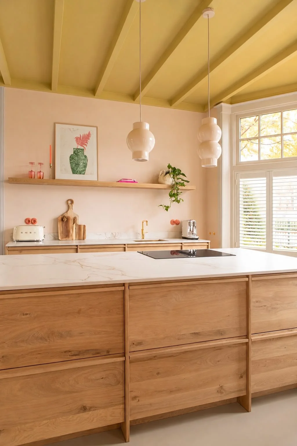 Modern kitchen with wooden cabinets, white marble countertop, and large window with shutters, decorated with plants and art.