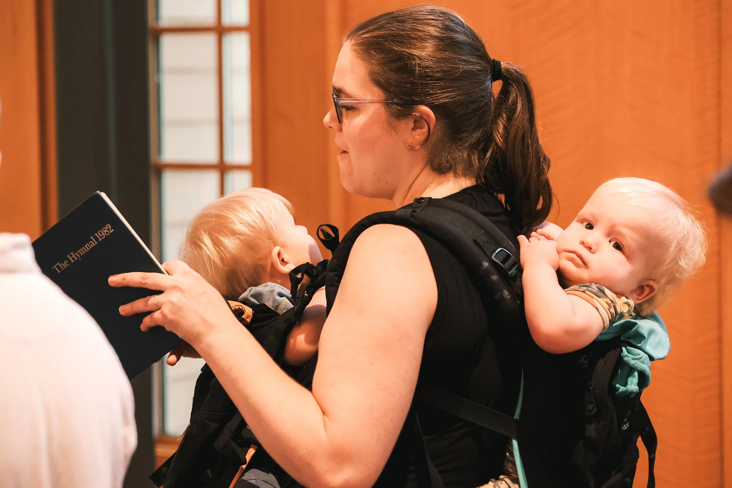 Episcopal Church of the Holy Cross: A woman standing and holding a hymnal in her hands singing with two small babies in carriers. One baby is being carried on her front and the other baby is being carried on her back