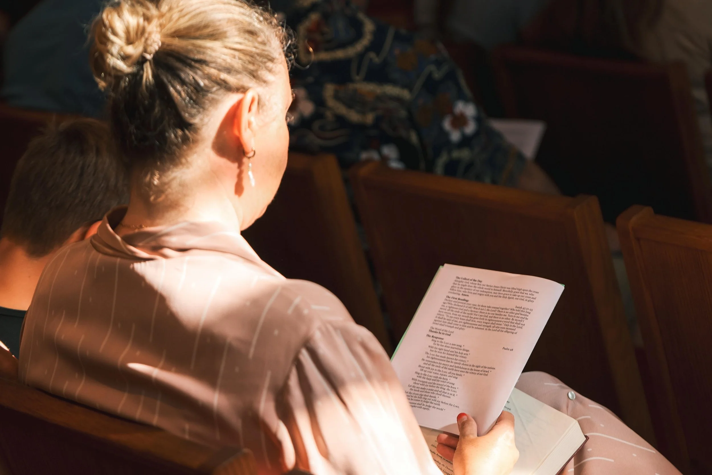 Holy Cross Episcopal Church: A woman with blonde hair tied in a bun and wearing earrings is reading a book while seated in a church pew.