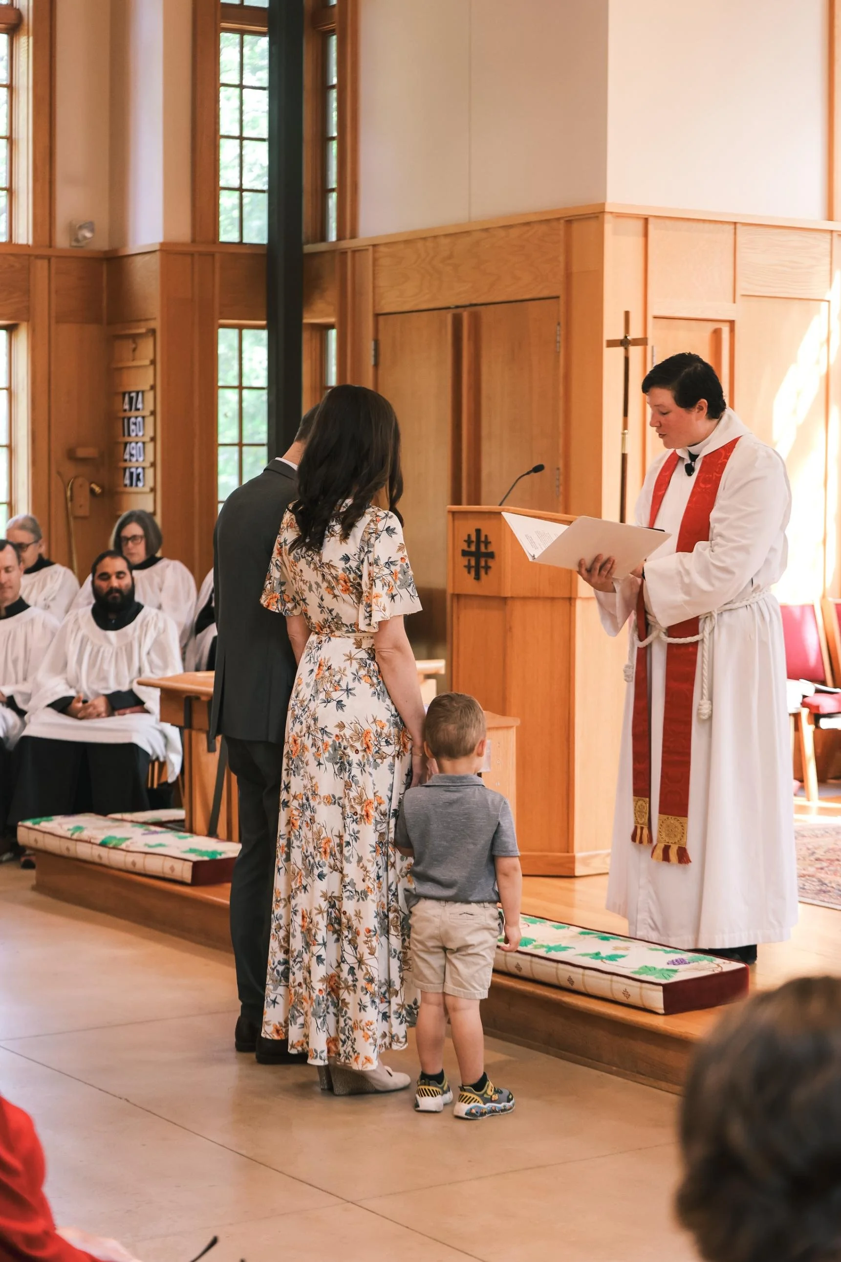Holy Cross Episcopal Church: A priest officiates a wedding ceremony inside a church. The bride and groom are standing in front of the altar, with two children also present. Several guests are seated in the background.