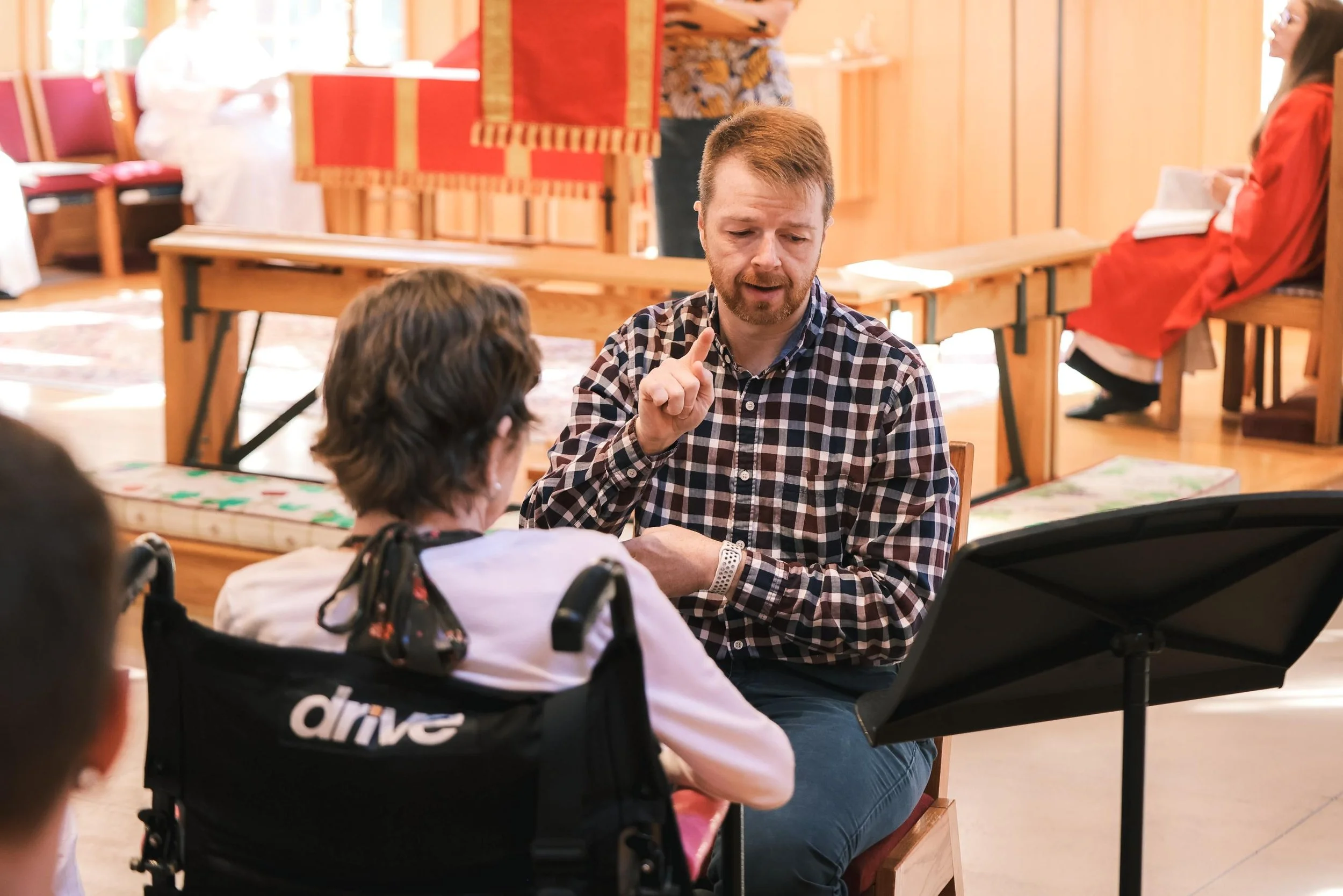Holy Cross Episcopal Church: A man with a beard and checkered shirt talking to a young girl in a wheelchair during a daytime indoor event with wooden decor and other people in the background.