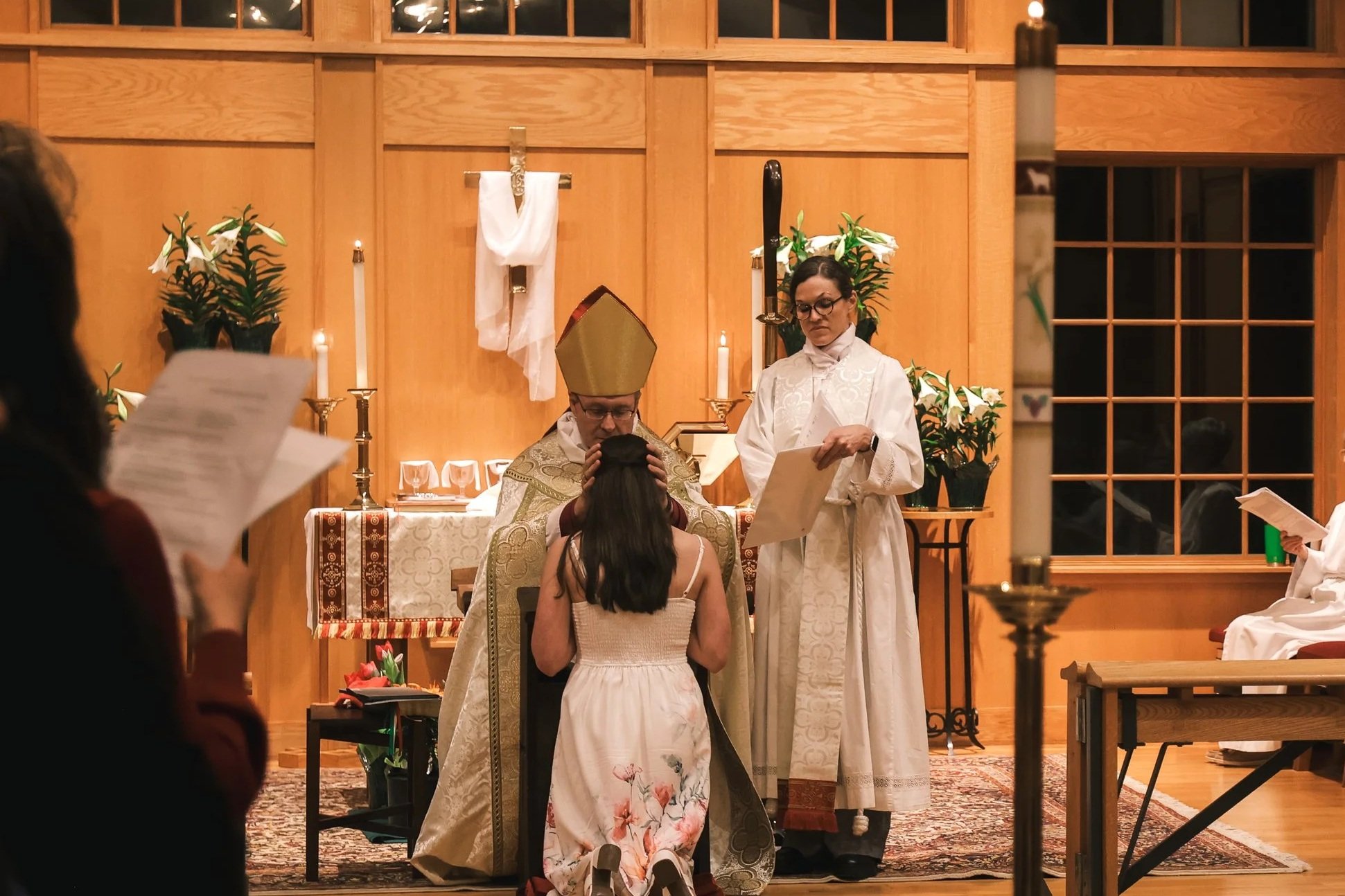 Holy Cross Episcopal Church: A religious ceremony inside a church with a bishop, a priest, and a woman kneeling before them, surrounded by lit candles and flowers.