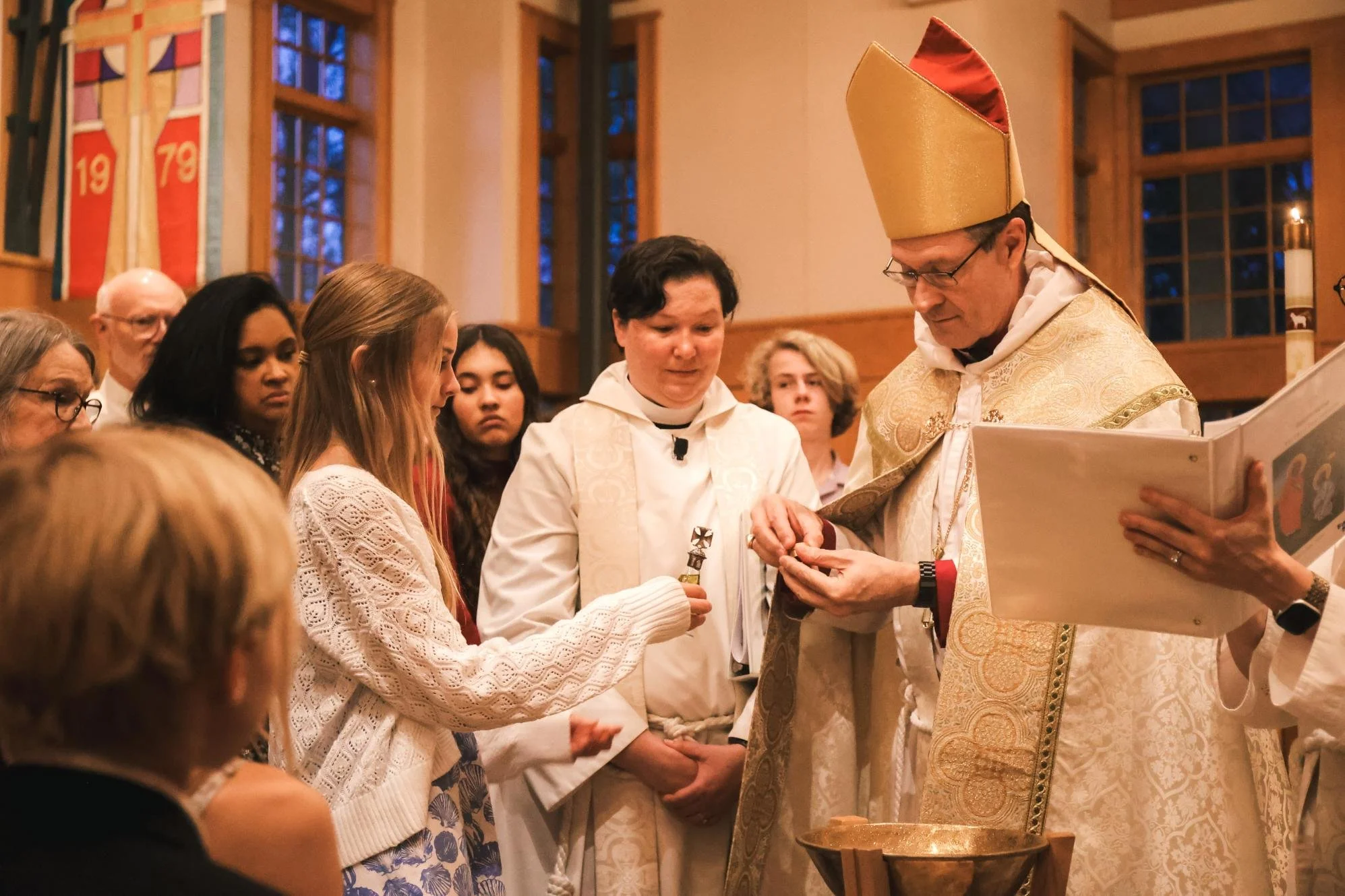 Holy Cross Episcopal Church: A religious ceremony in a church where a bishop, wearing a gold mitre and ornate vestments, is receiving a gift of baptismal oil from a teenage girl in a white sweater.