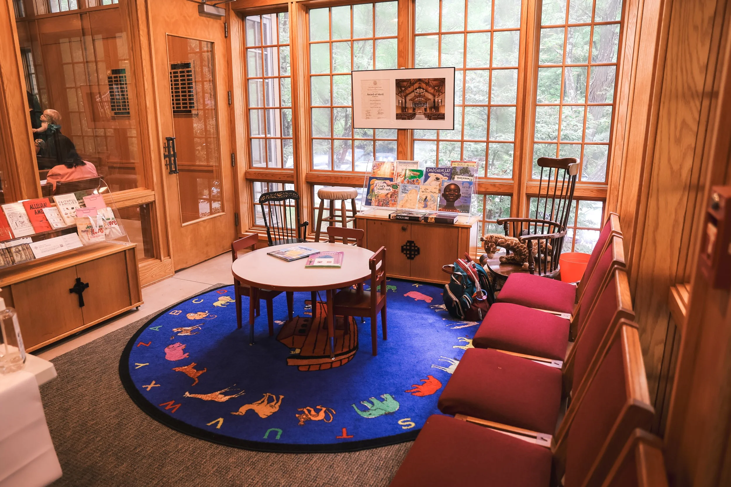 Episcopal Church of the Holy Cross: A welcoming and friendly space designed for children surrounded by glass windows. There is a small table, rocking chair, stuffed animals, and children's books.