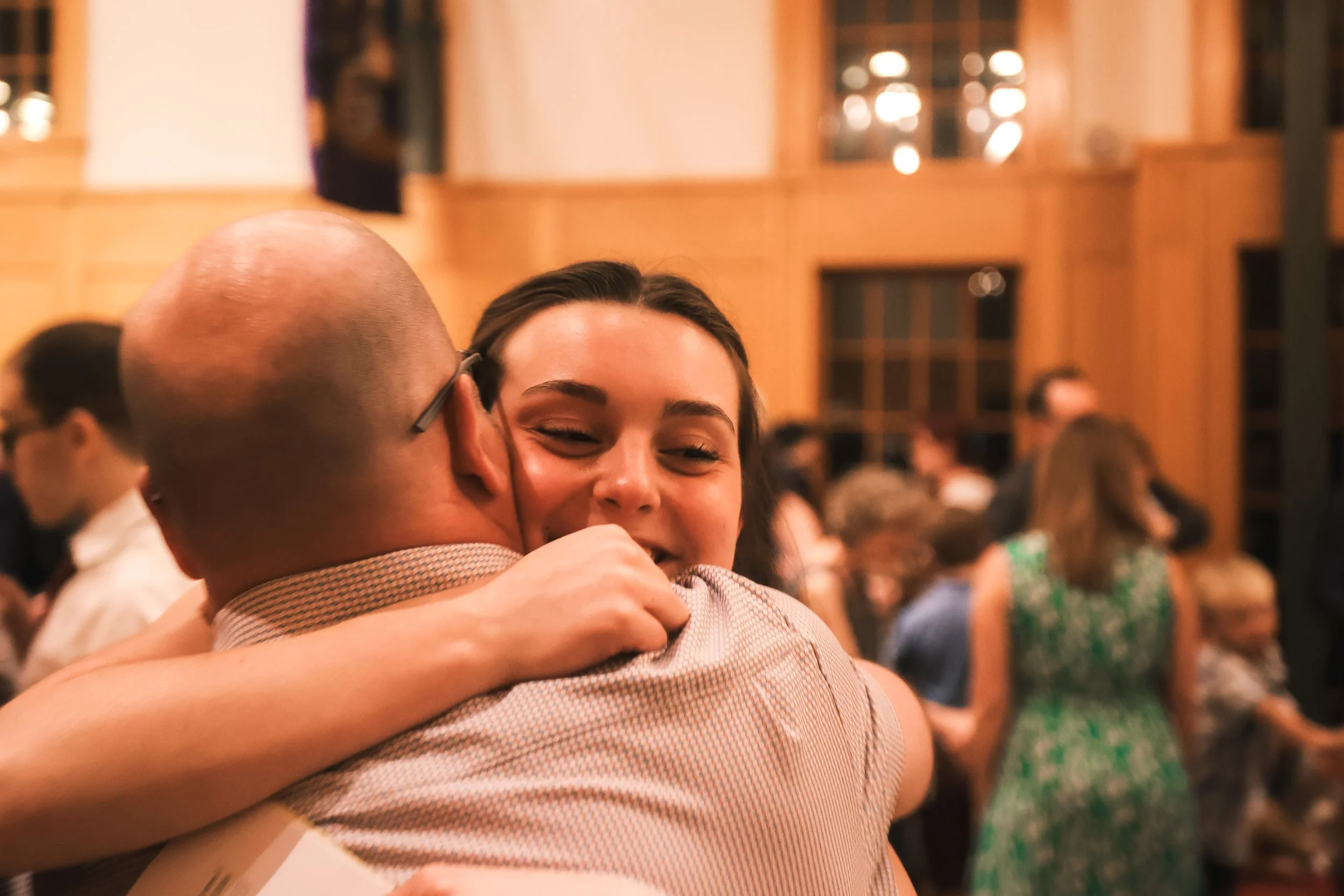Holy Cross Episcopal Church: A man and a woman are hugging, both with joyful expressions, at a social gathering or celebration with several people in the background.