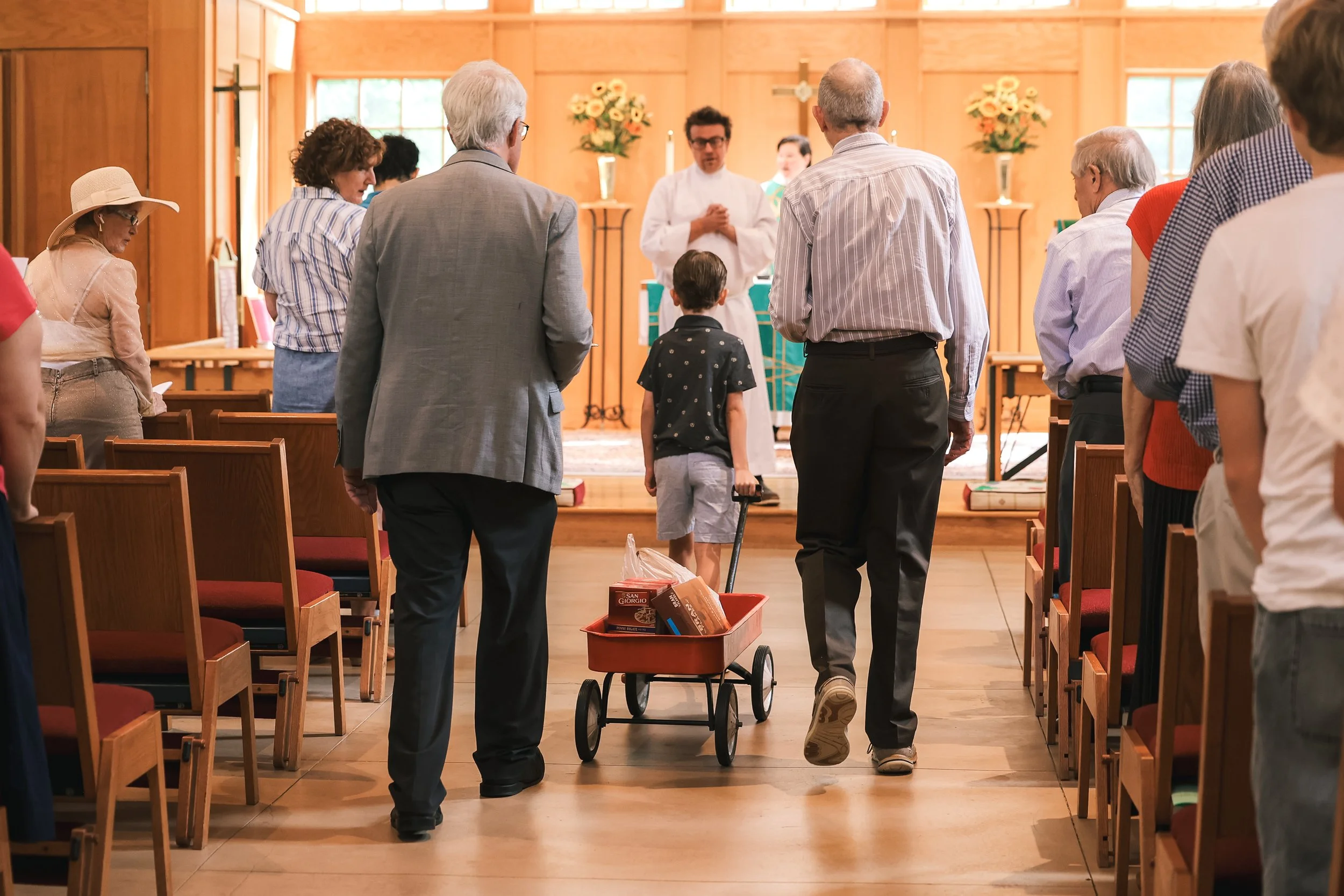 Episcopal Church of the Holy Cross: A religious ceremony where a young boy in a navy shirt pulls a red wagon full of food for those in need toward the altar.