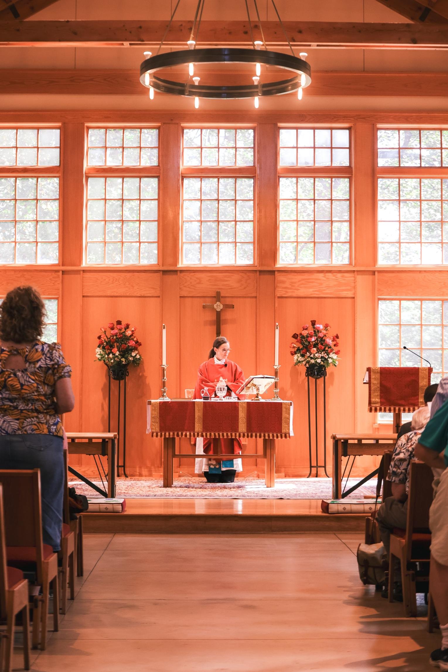 Holy Cross Episcopal Church: Interior of a church or chapel with a wooden altar, a large cross at the center, and a woman in a red robe standing behind the altar, singing or speaking. There are flowers, candlesticks, and a lectern on the altar.