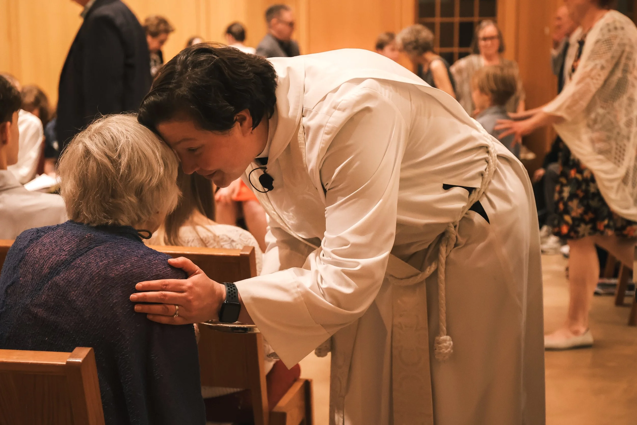 Holy Cross Episcopal Church: A man in a white shirt is bending down to bow and place a hand on an elderly woman's shoulder during a religious or cultural ceremony inside a warmly lit room with wooden paneling.
