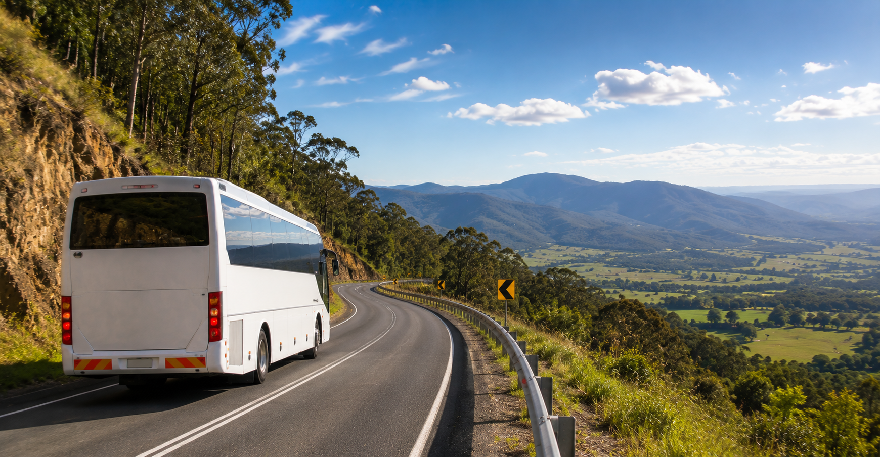 A white bus driving on a winding mountain road with guardrails, surrounded by green trees and hills, under a partly cloudy blue sky.