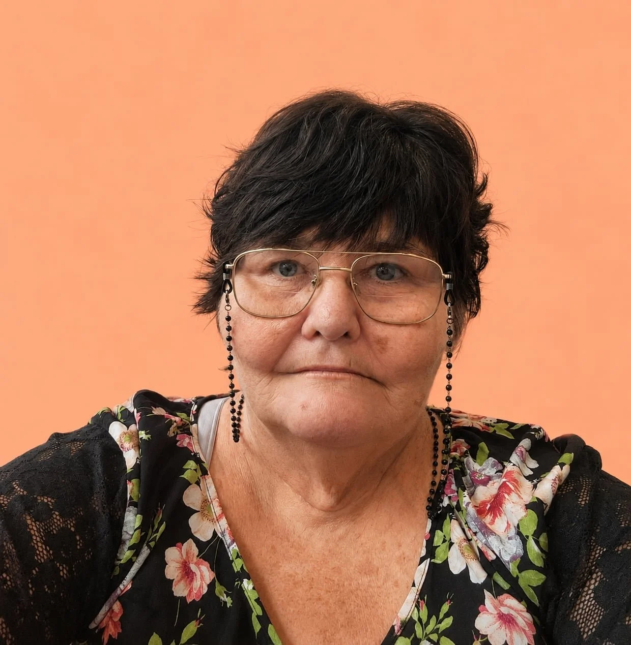 Close-up of an older woman with short dark hair, wearing large glasses with black bead chains, earrings, and a floral top, set against a peach-colored background.