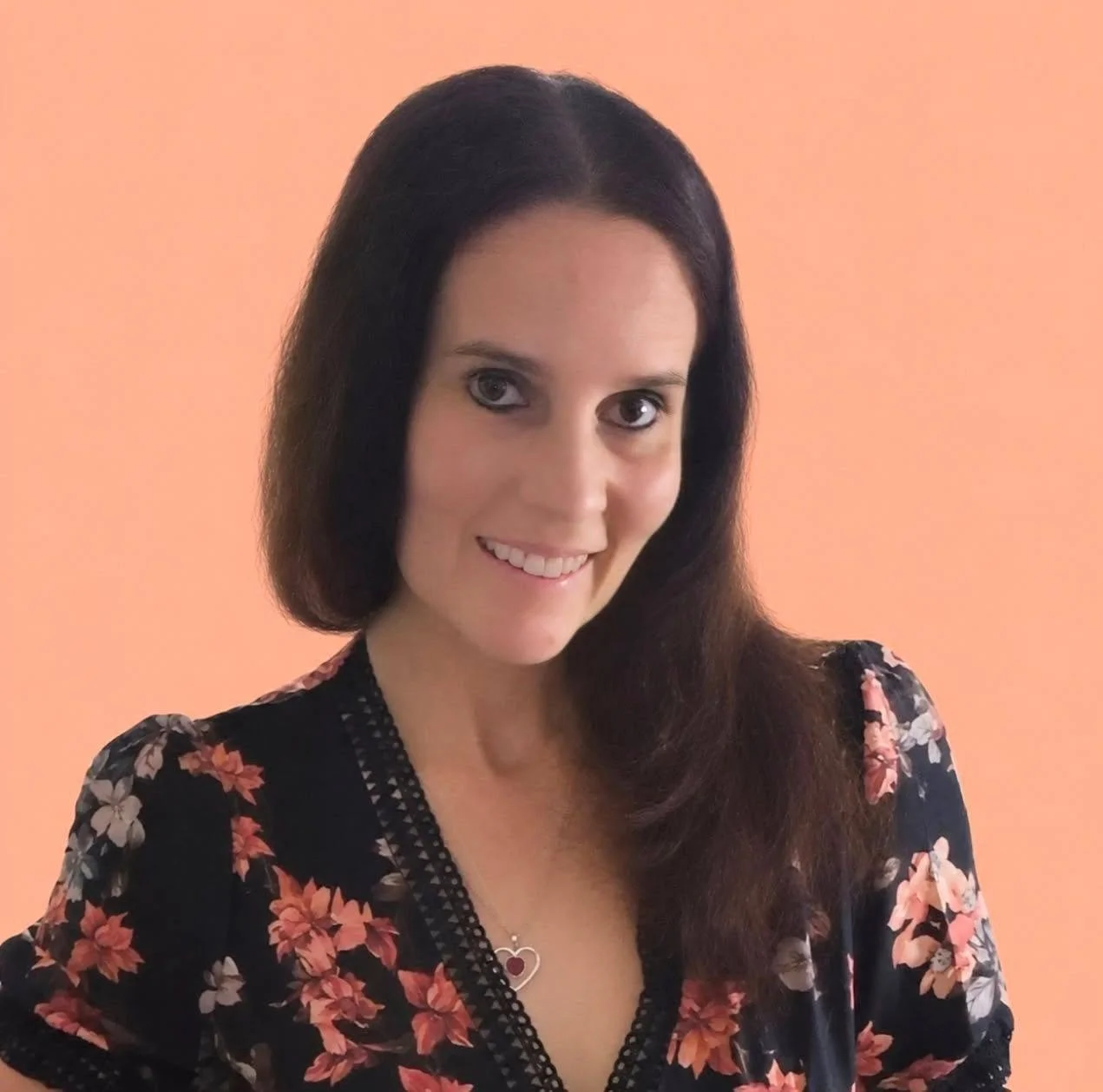 A woman with long dark hair, light skin, and blue eyes, smiling, wearing a floral black dress and a silver necklace with a heart-shaped pendant, against a peach-colored background.
