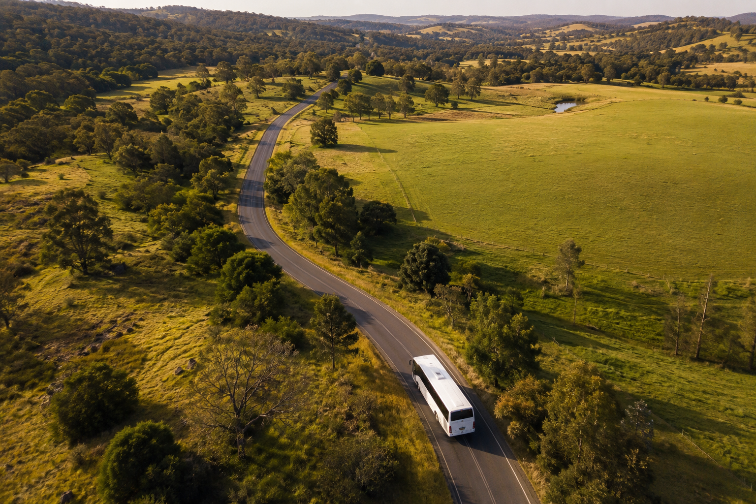 Aerial view of a winding two-lane highway through green countryside with trees and grassy fields, a white bus traveling on the road, and hills in the distance.