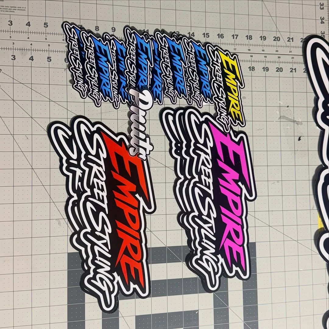 Multiple colorful stickers with the text 'EMPIRE', 'S' and 'STREETSUNGS' arranged on a grid cutting mat.