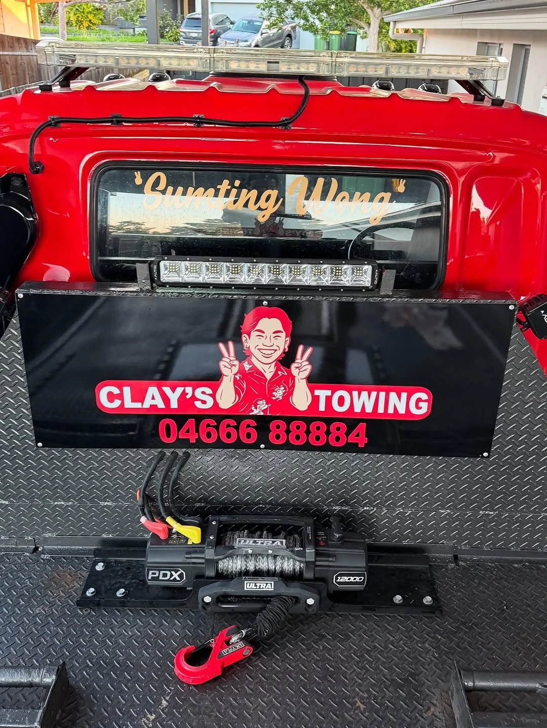 The back of a red tow truck with a black panel that displays the name 'Clay's Towing' and a cartoon image of a smiling man with red hair making peace signs. The unit also has a LED light bar mounted on top, and there is a winch with a hook on the flo