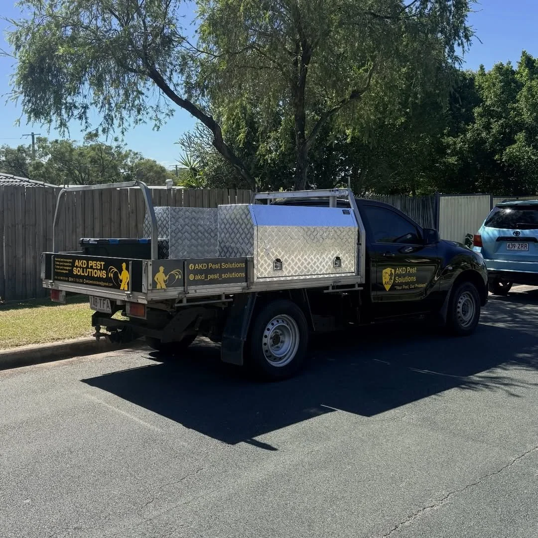 A black work truck with yellow signage for AKD Pest Solutions parked on the side of a suburban street. The truck has a flatbed with metal storage compartments and a fence around the back. There are trees and a wooden fence in the background.