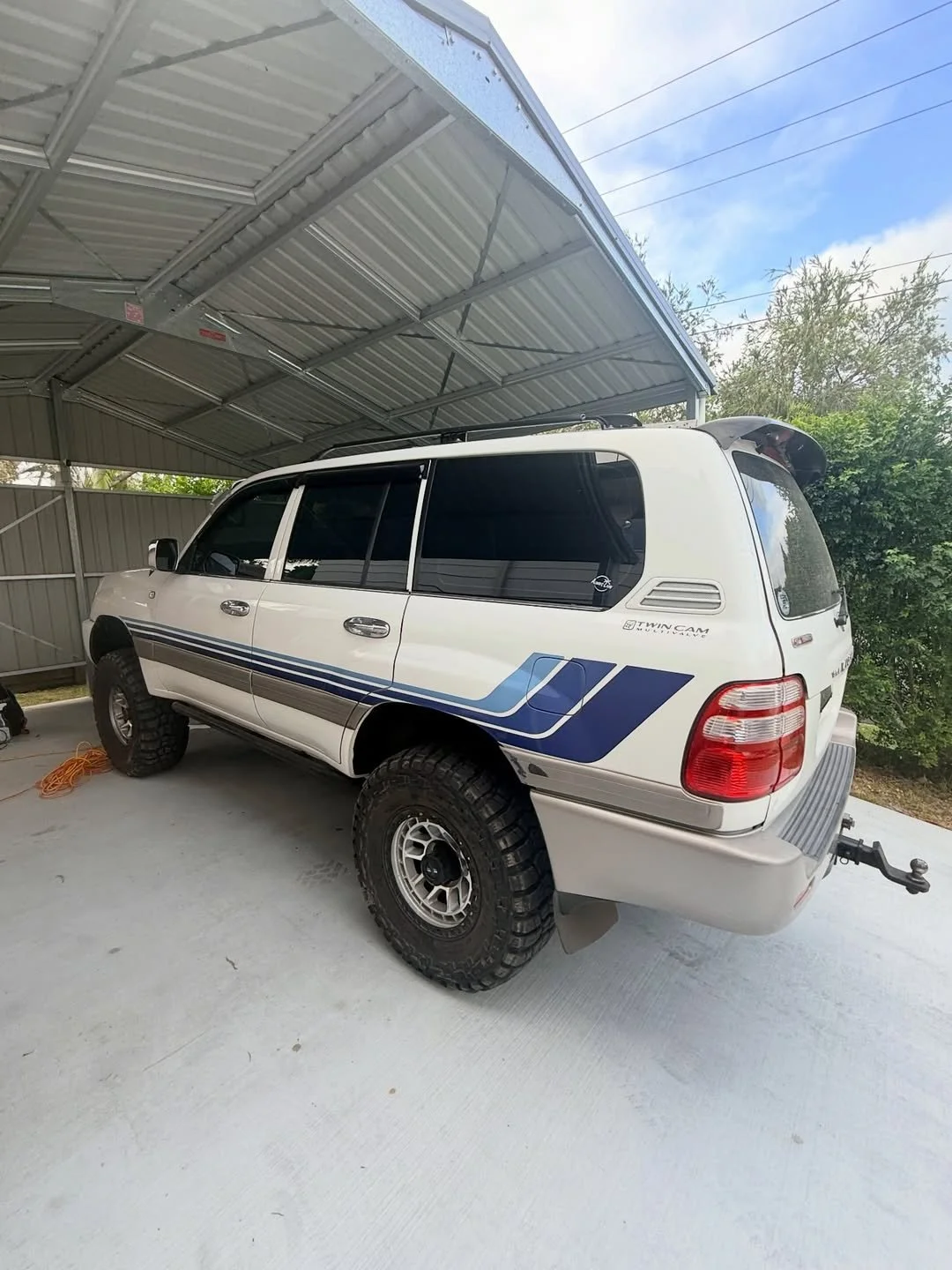 A white SUV with large off-road tires parked under a metal carport, with a sticker on the rear side window indicating it has a twin cam engine and a trailer hitch attached.