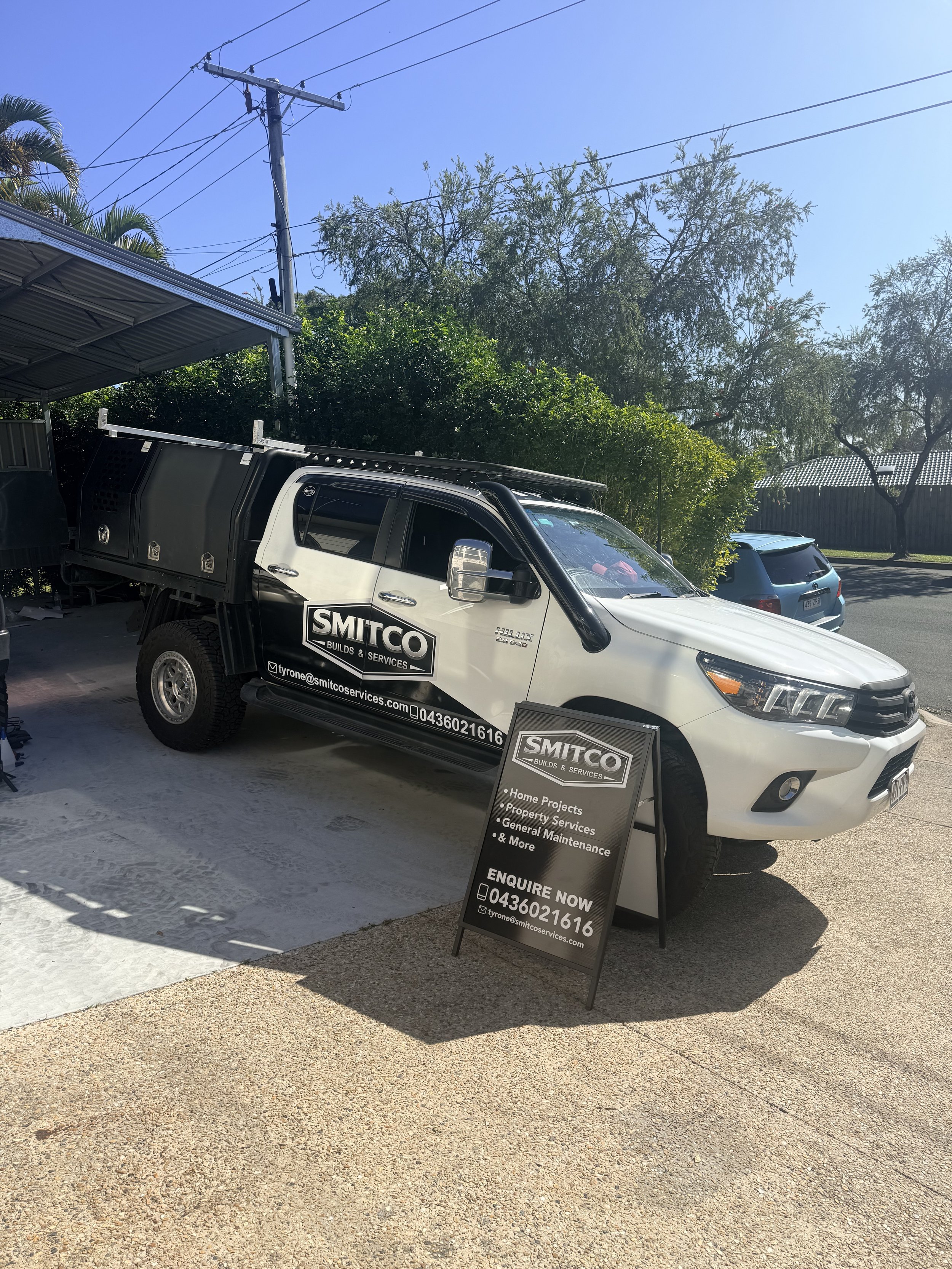 A white pickup truck with black signage for Smitco Builds & Services parked under a metal carport, with a black sandwich board sign in front advertising home projects, property services, general maintenance, and more, on a sunny day with blue sky and